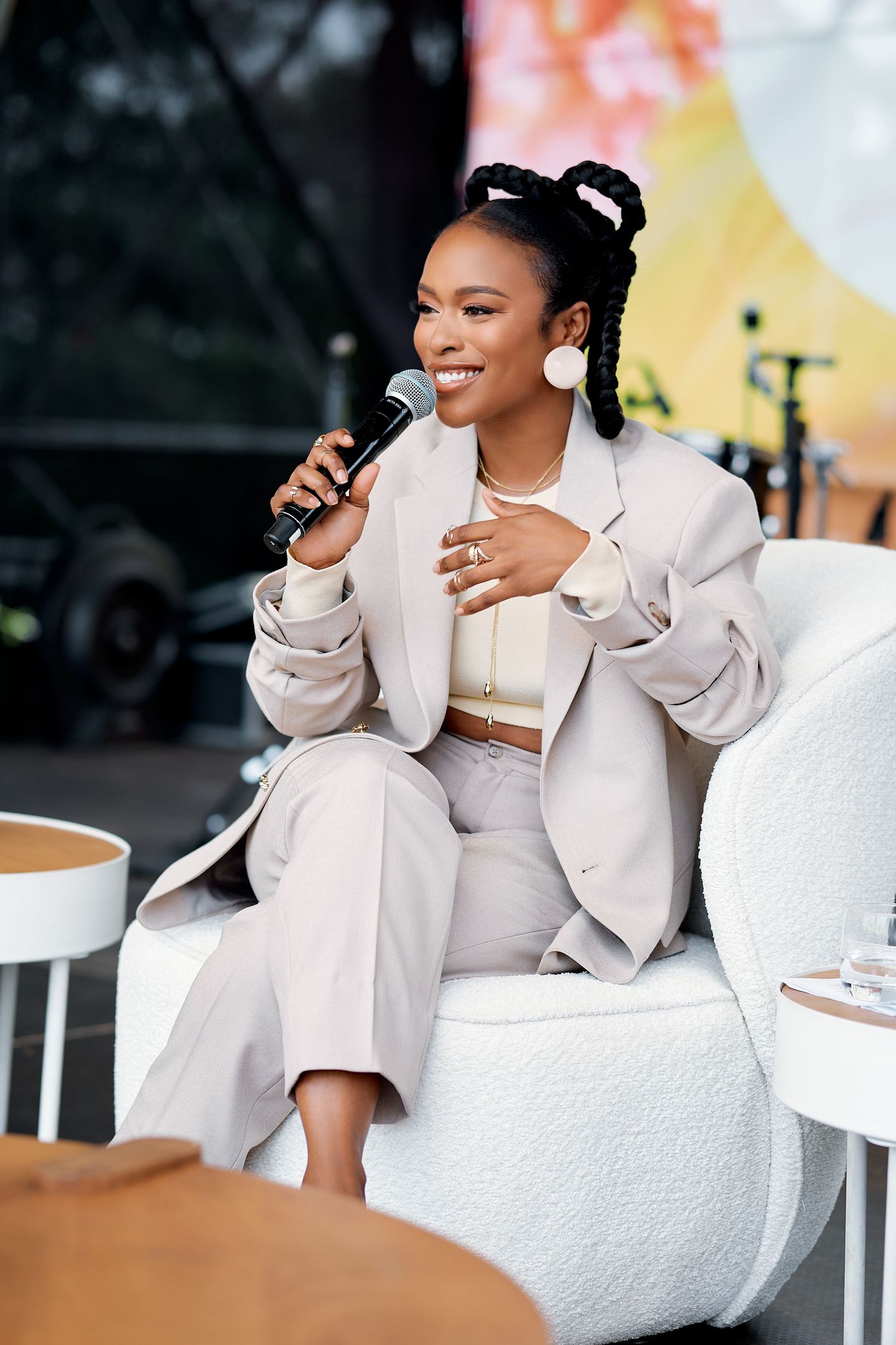 Smiling woman with braids holding a microphone while seated at an outdoor speaking event.