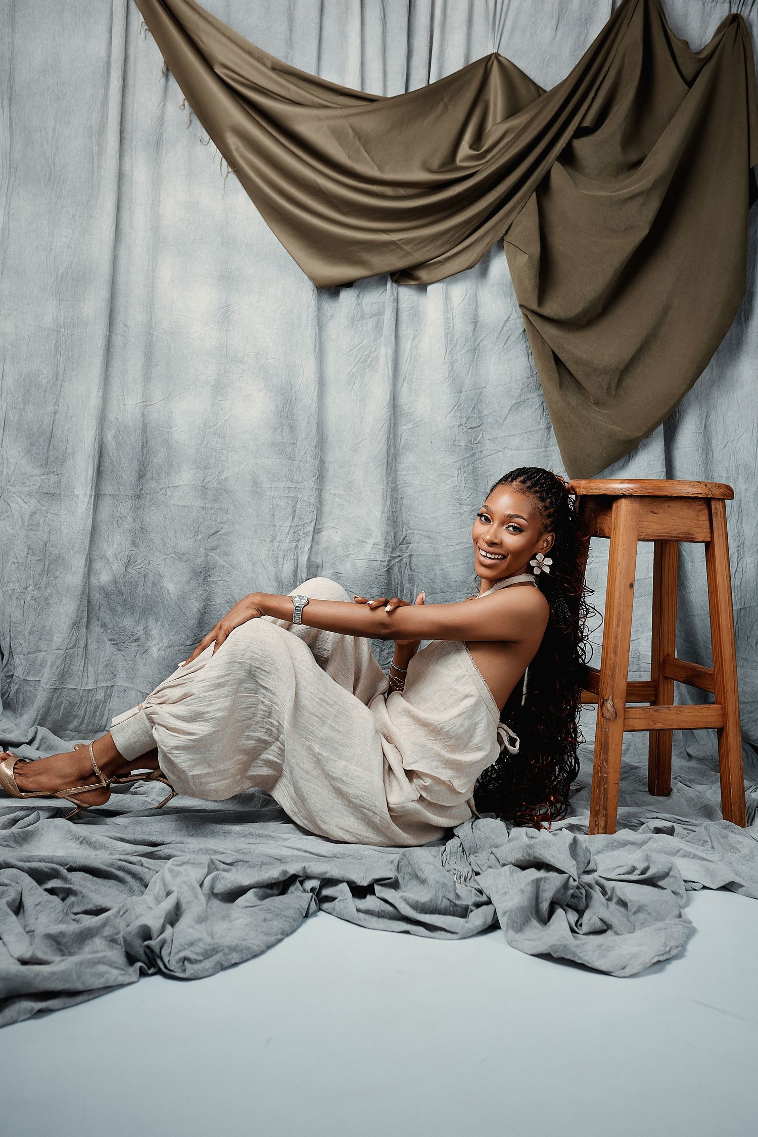 Smiling woman with braids in beige outfit sitting on floor beside wooden stool in studio.
