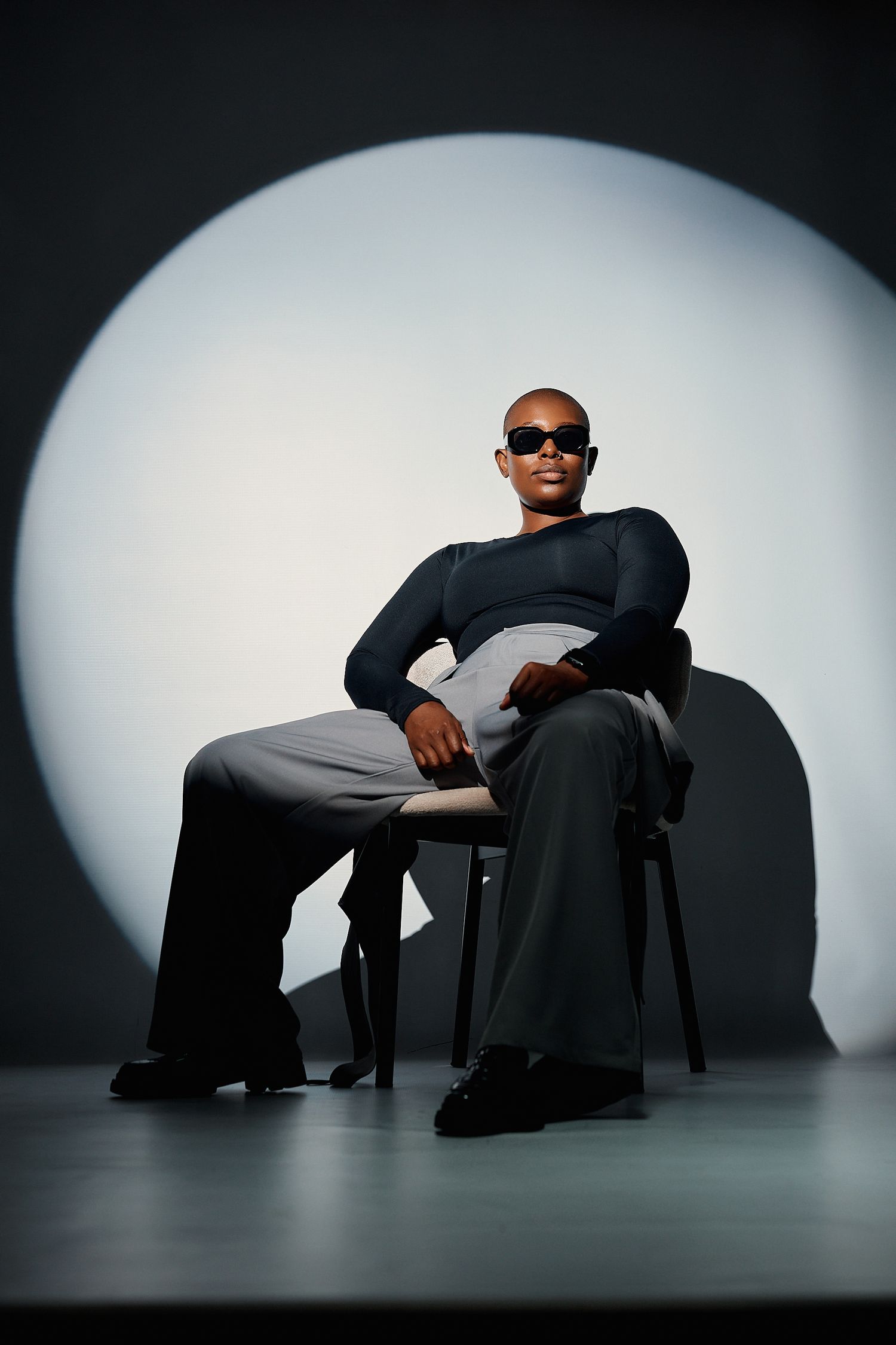 Bald Black man in dark fitted top and gray trousers sits confidently on chair wearing sunglasses against circular light backdrop.
