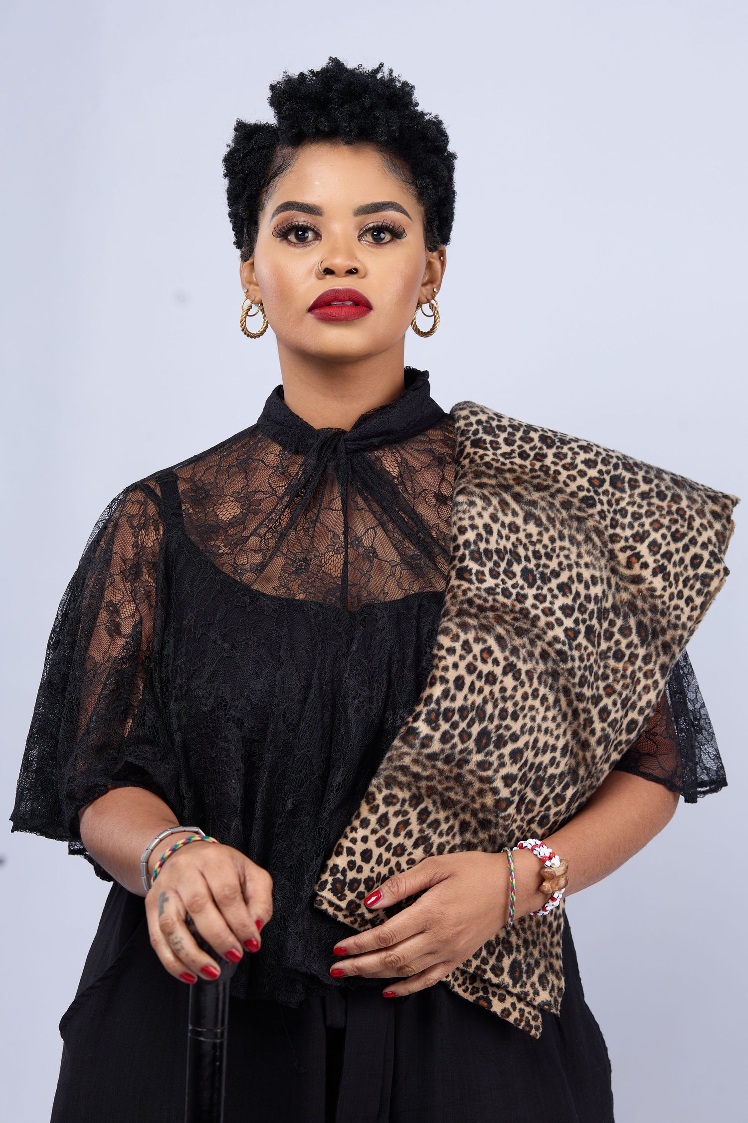 Woman in black lace top holding leopard print clutch, wearing gold hoop earrings and red lipstick, studio portrait.