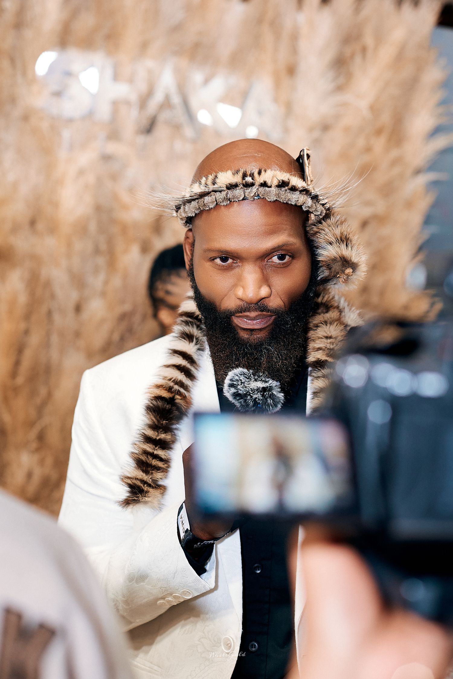 Bald Black man with a beard wearing traditional African fur attire being filmed at an event.
