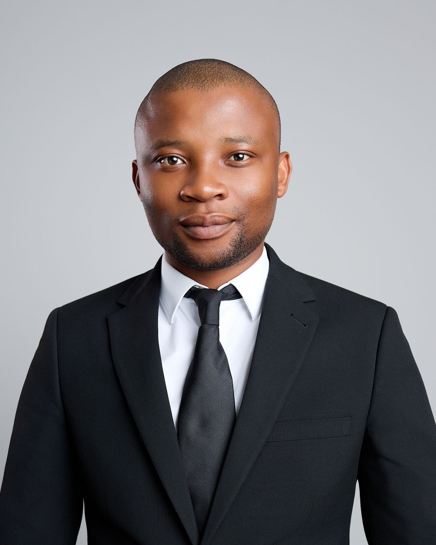 Young Black man in black suit and tie poses for professional headshot on gray background.