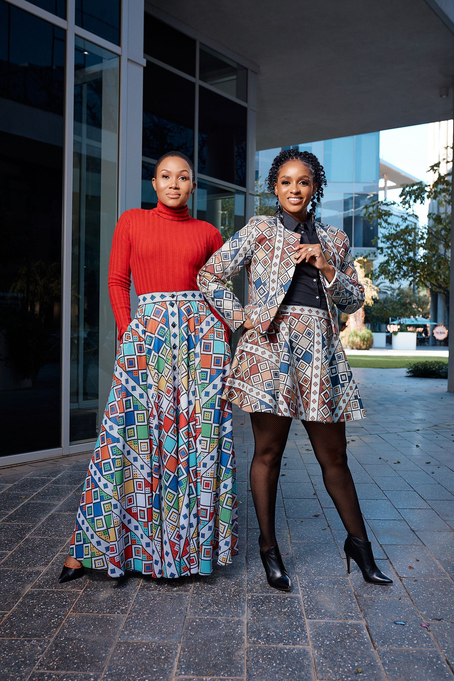 Two women wearing African print skirts and jacket posing together outside glass building.