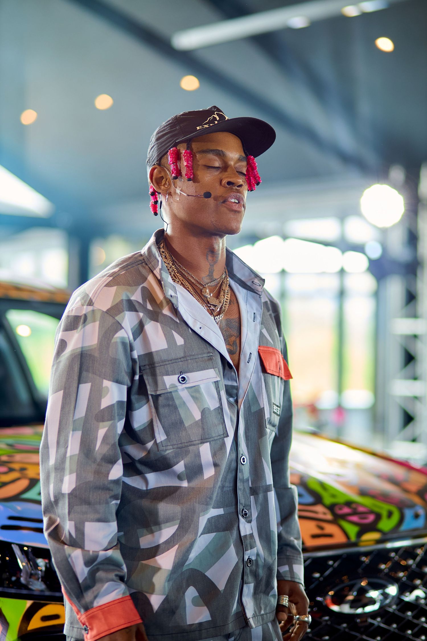 Young man with pink braids in camo jacket posing beside colorful painted car indoors.