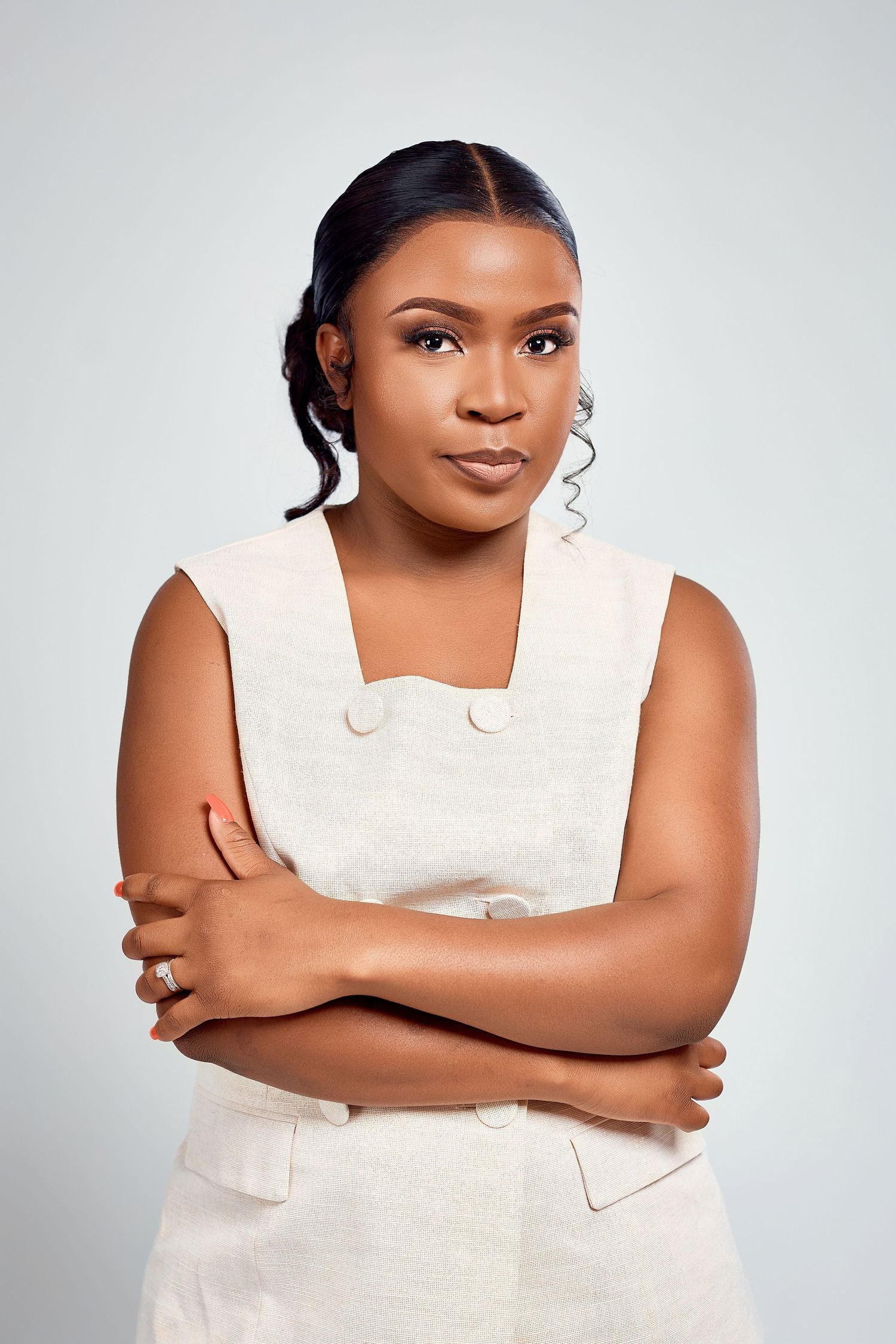 A confident young Black woman in a cream sleeveless dress stands with arms crossed against a light gray background.