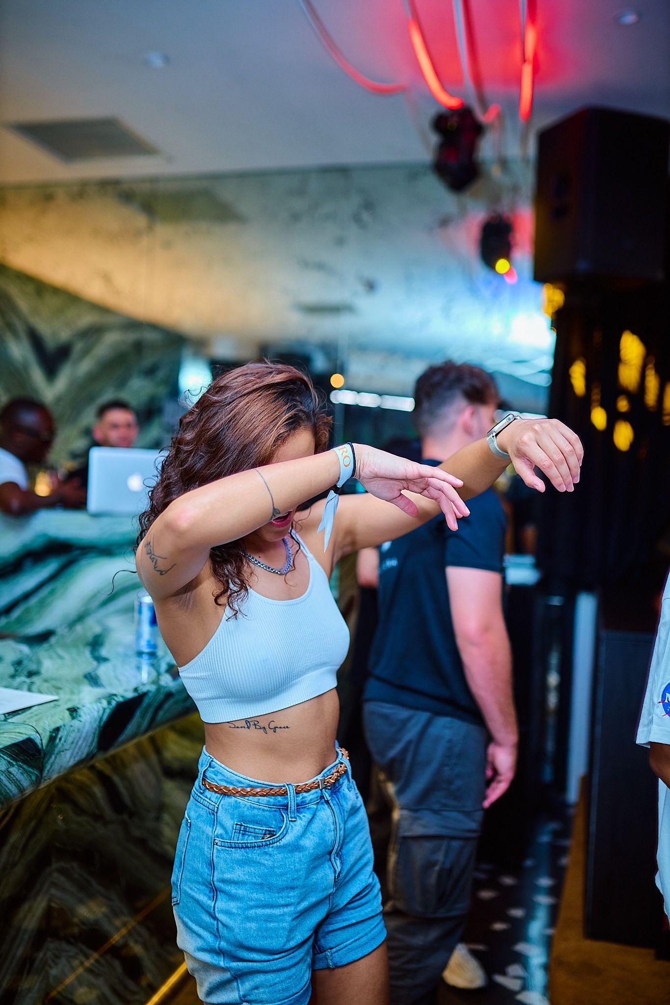 Young woman dancing in a white crop top and denim shorts at a vibrant nightclub or bar event.