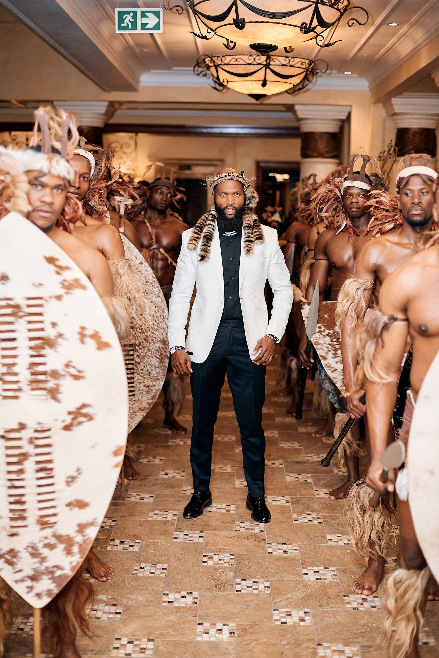 Man in white blazer and black suit stands between rows of traditional African warriors holding shields in elegant venue.