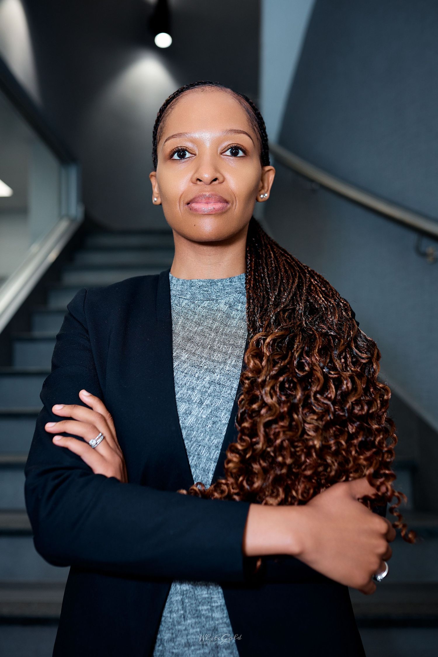 Professional Black woman with long braided hair stands arms crossed near staircase in modern building.