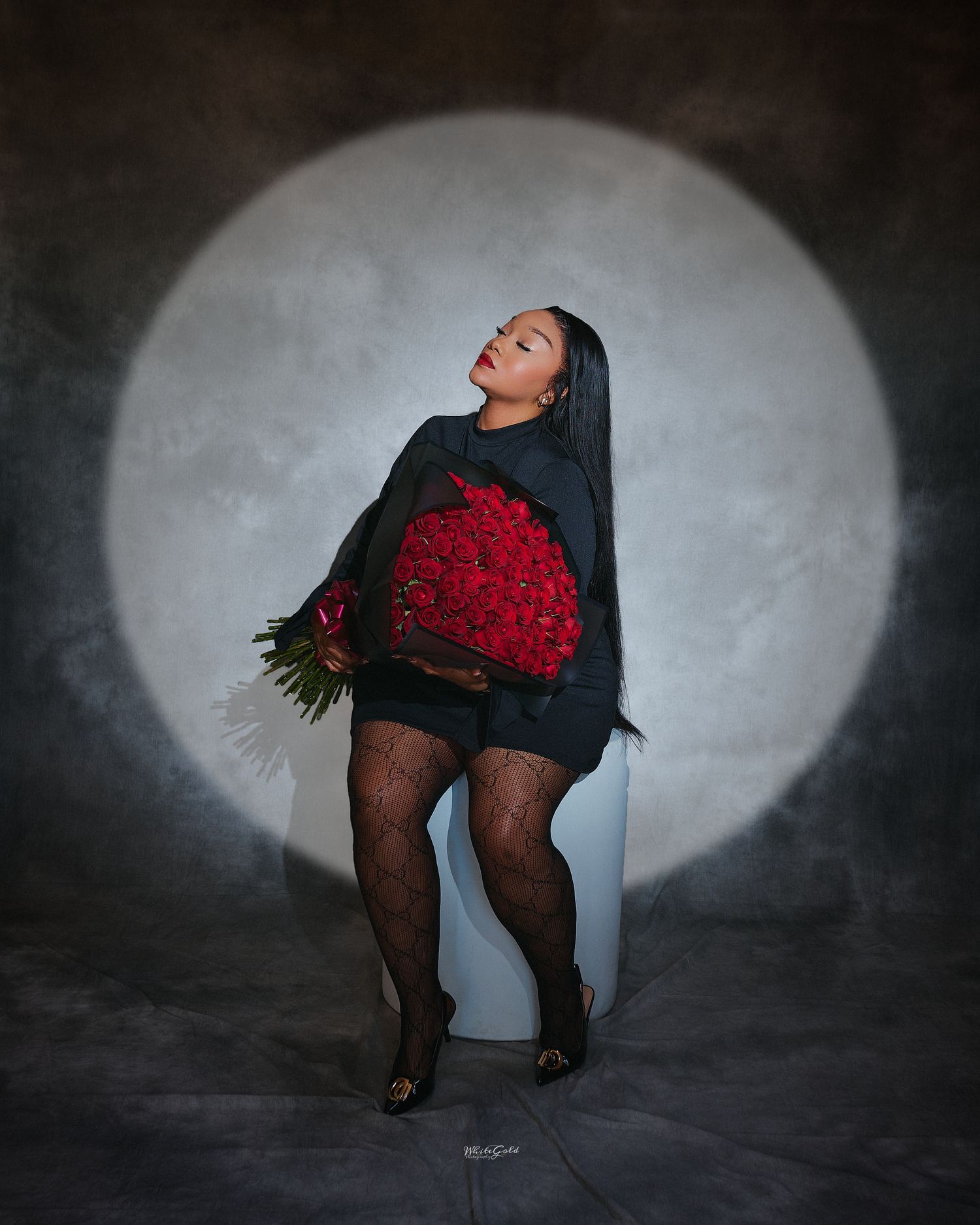 Elegant Black woman poses with large red rose bouquet in black outfit against dramatic spotlight backdrop.