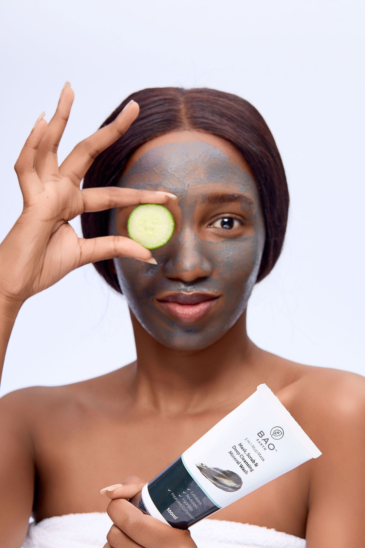 Woman with clay face mask holding cucumber slice over eye and skincare tube product.