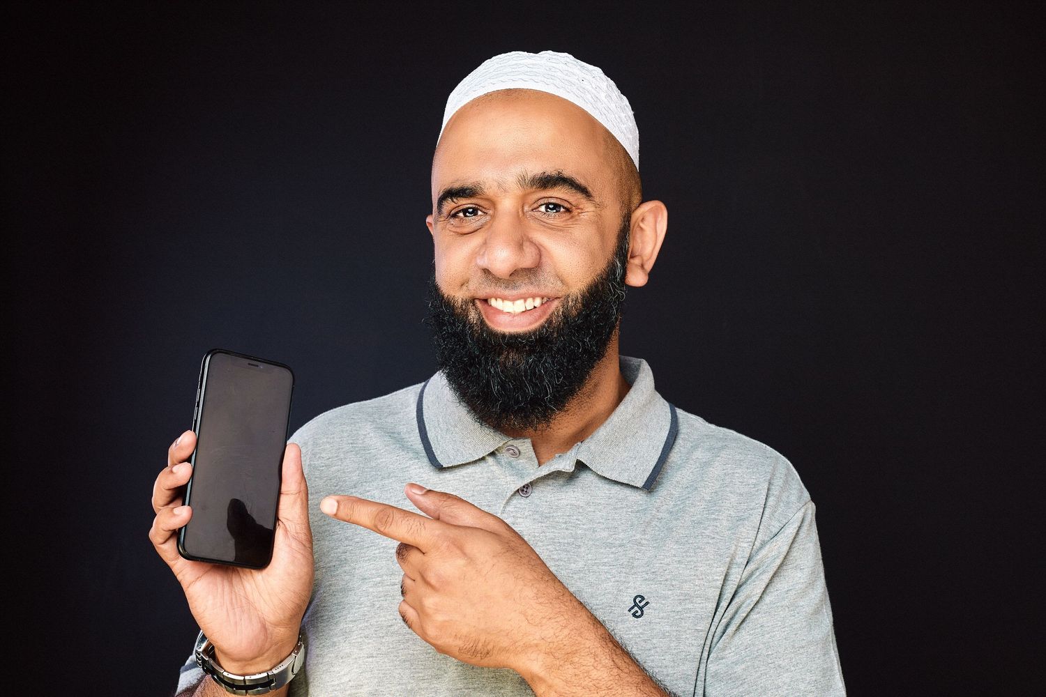 Smiling bearded Muslim man in gray polo and white kufi pointing at a smartphone on dark background.