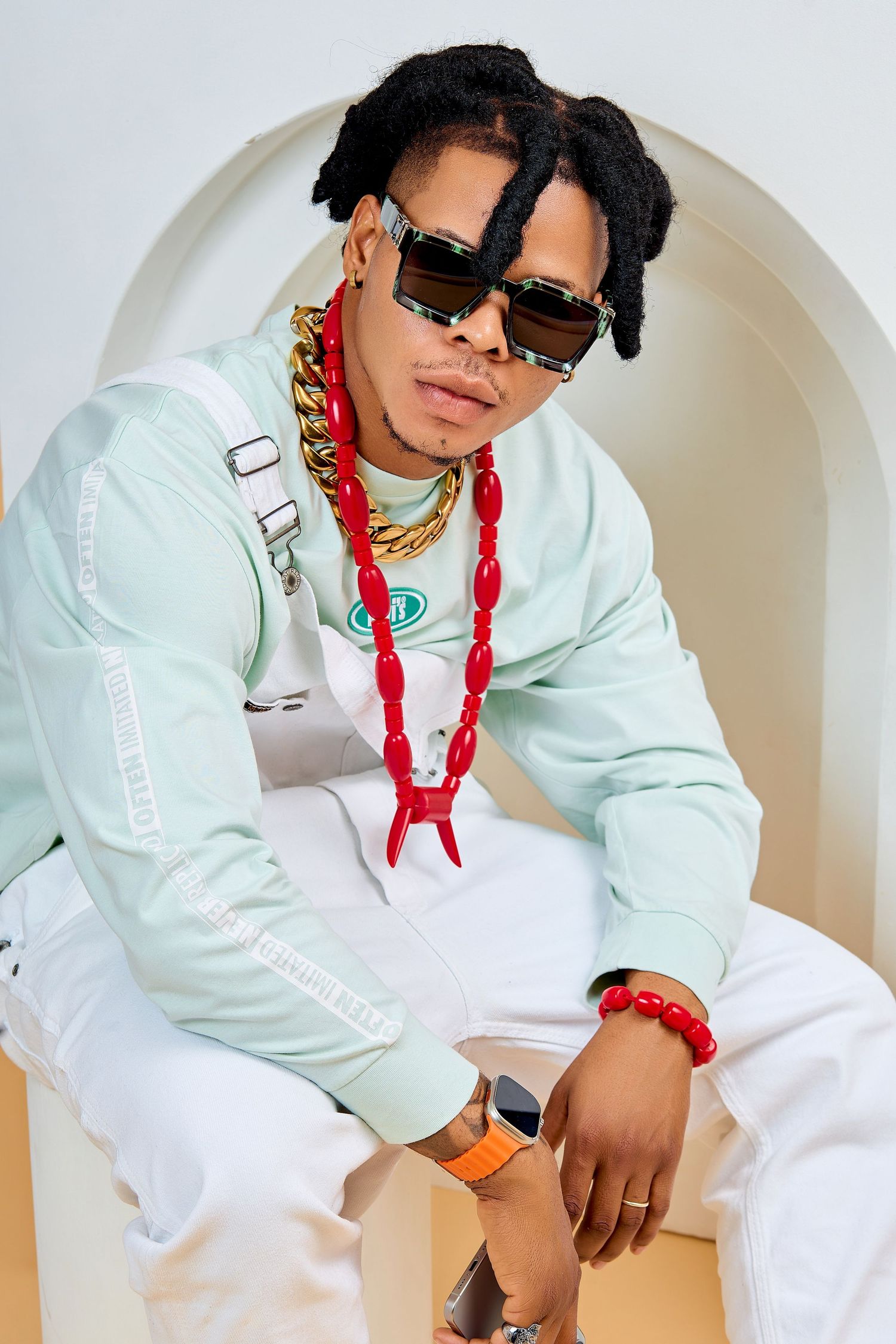 Trendy Black male artist with dreadlocks wearing mint tracksuit and layered gold and red chains sits posing.