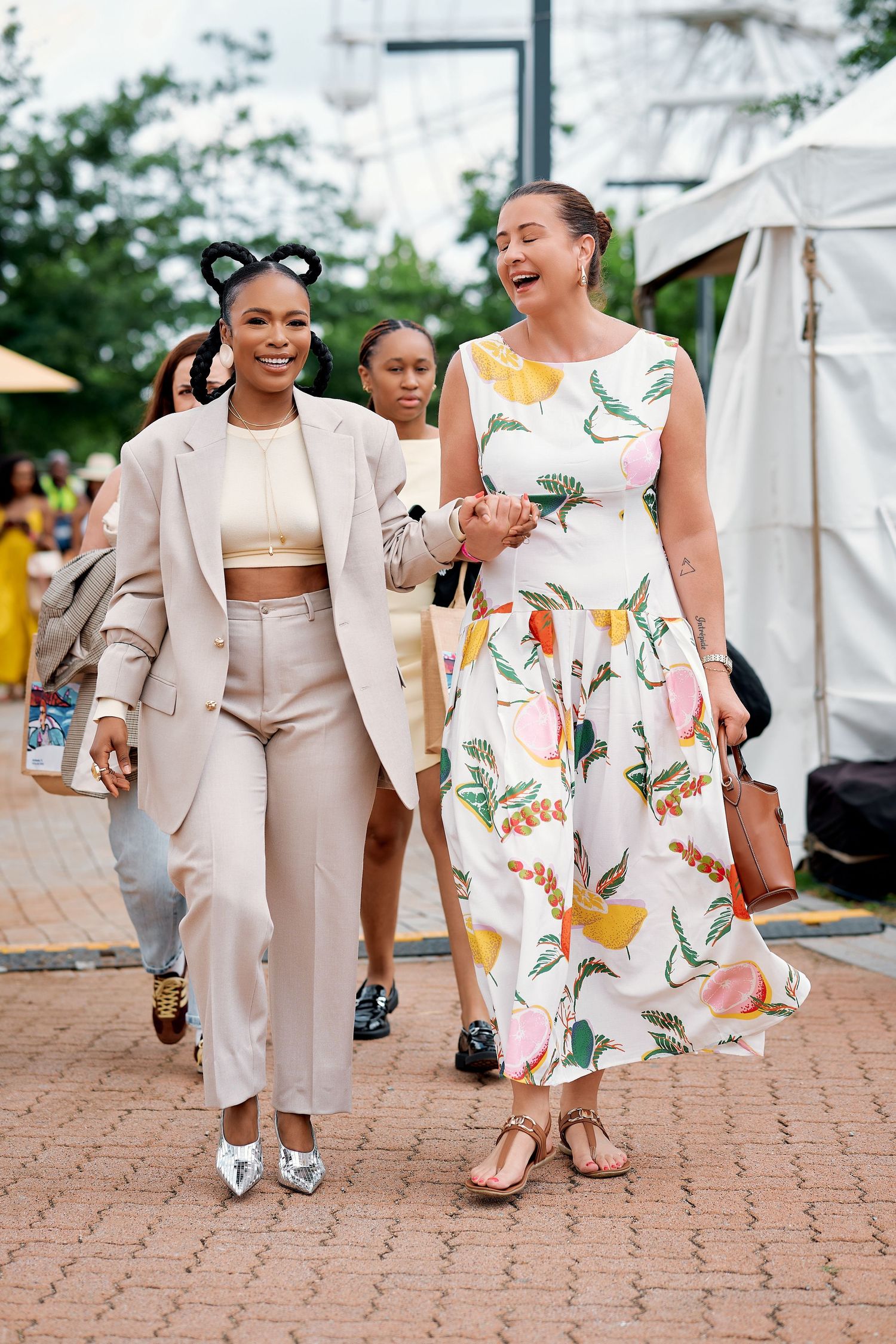 Two stylish women walk and laugh together at an outdoor summer fashion event or festival.