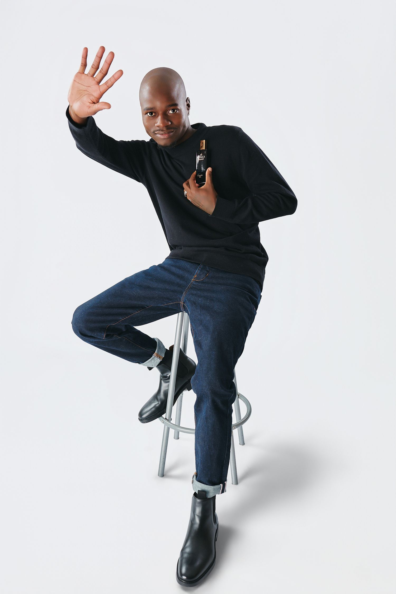 Bald man in black sweater and jeans sitting on stool holding perfume bottle while waving.