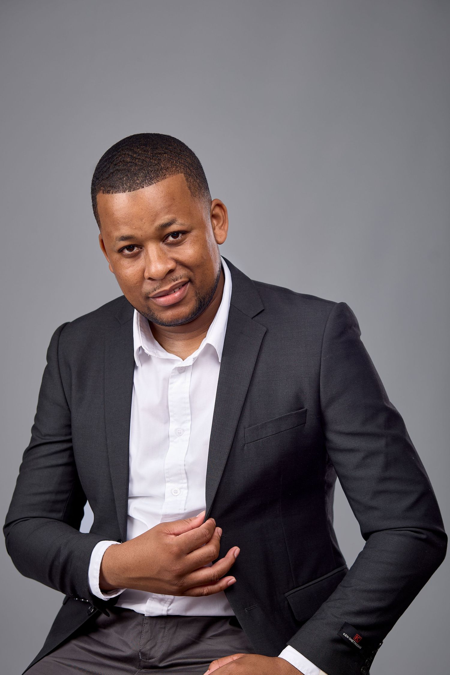 Professional Black man in dark suit and white shirt poses confidently against gray background.