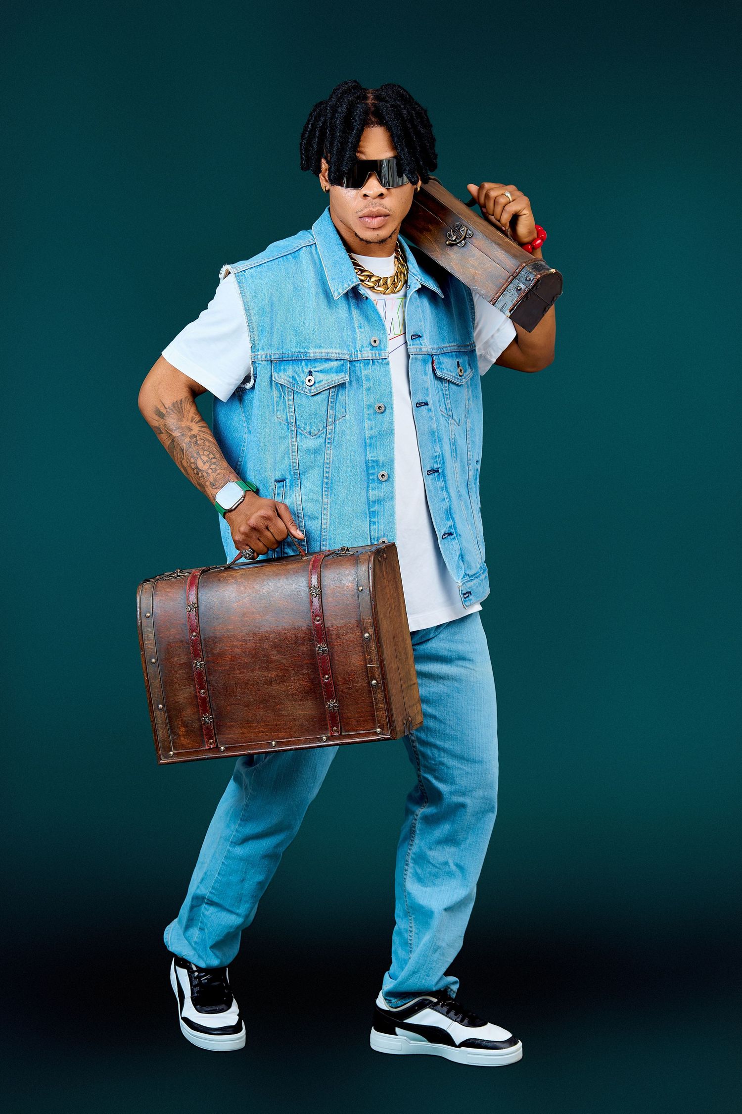 Cool Black male artist with dreadlocks in denim vest carries vintage suitcase and boombox on teal background.