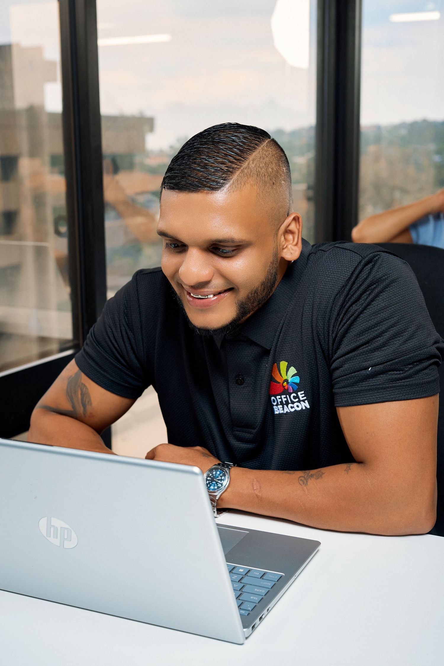 Smiling young man in an Office Beacon polo shirt working on an HP laptop in a modern office.