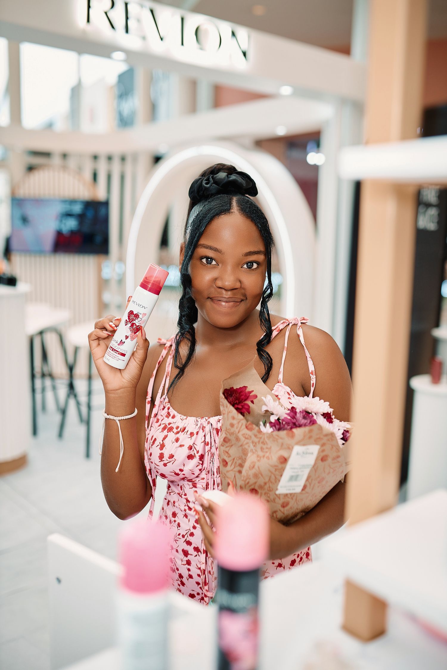 Smiling woman in floral dress holding Revlon beauty product and flower bouquet at Revlon store.
