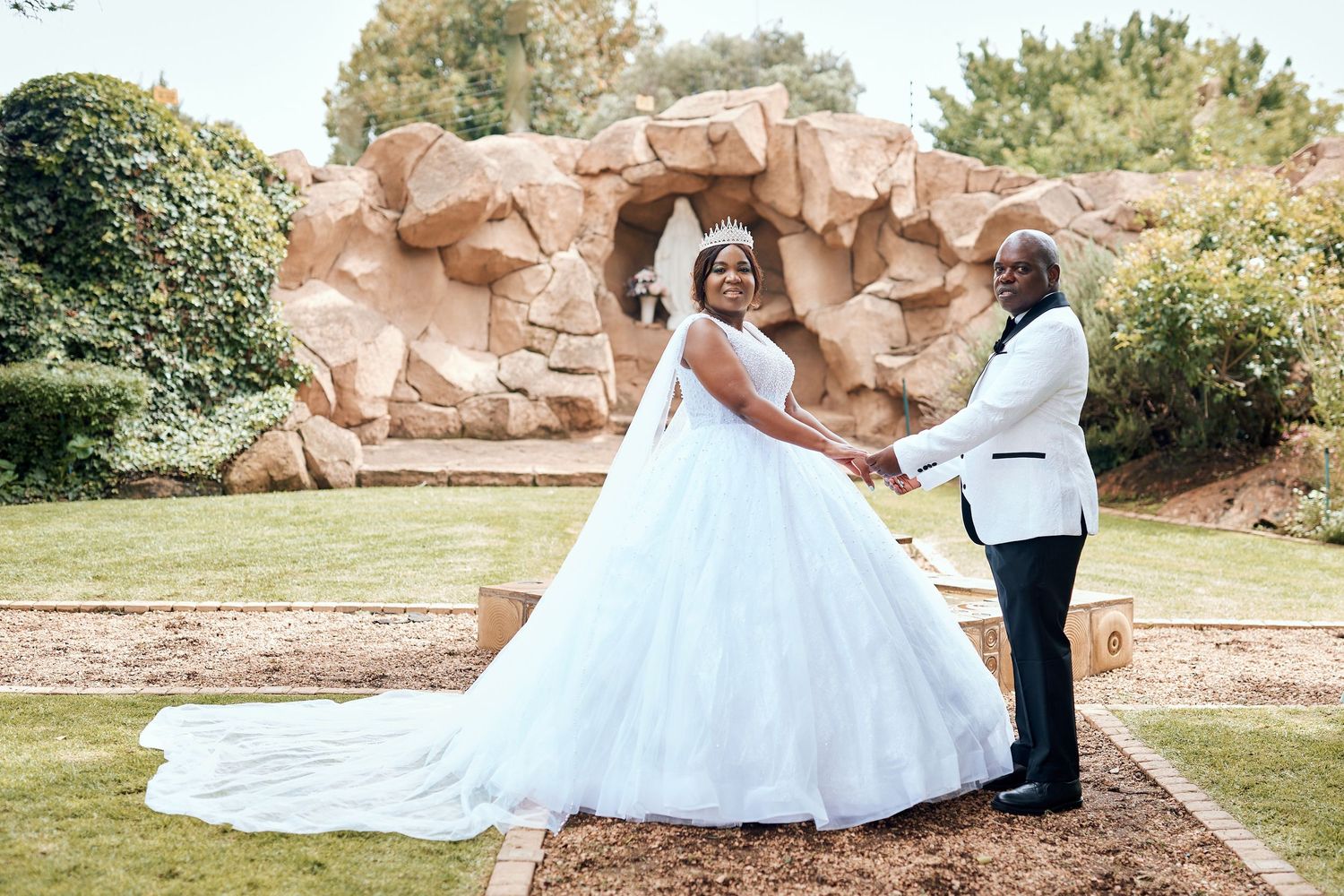 Happy Black couple in wedding attire posing before a stone arch in a lush garden setting.