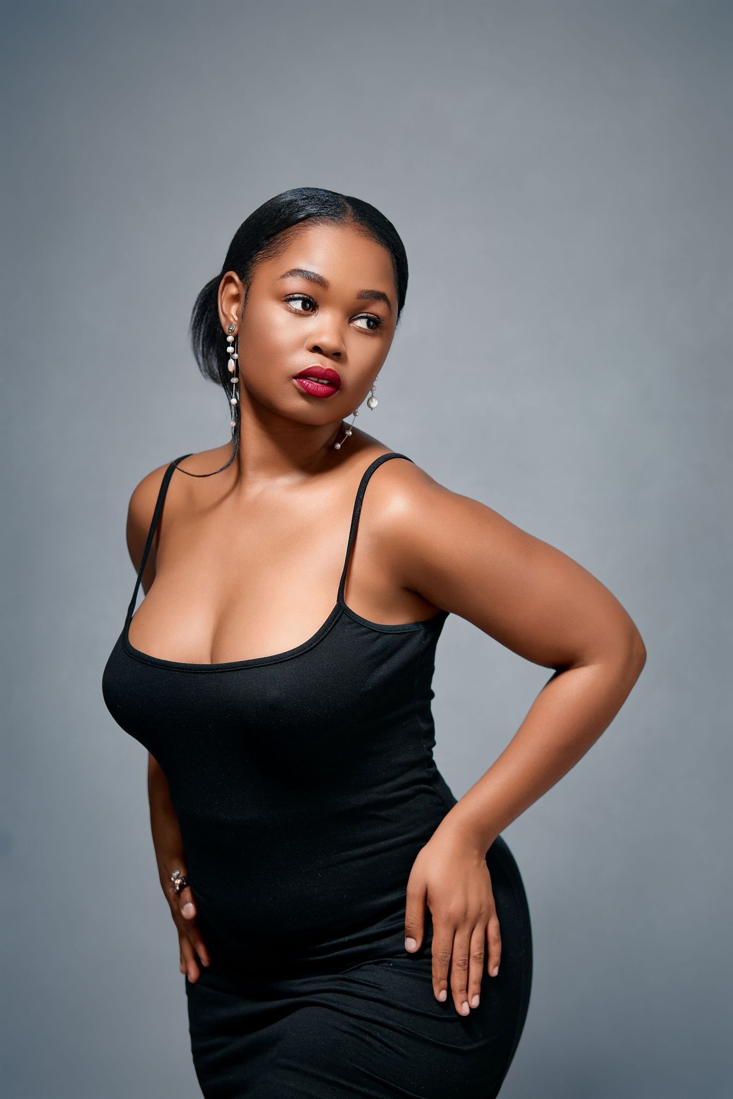 Stylish Black woman in fitted black dress with dangling earrings poses gracefully against gray background.