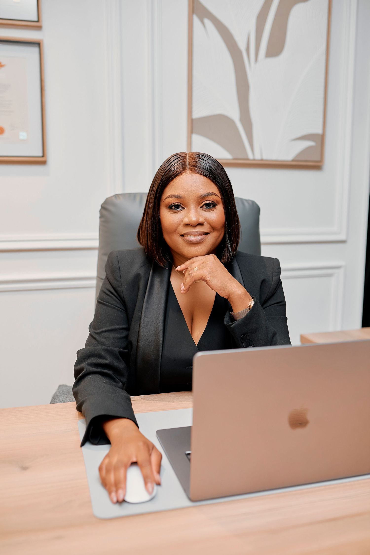 Confident Black businesswoman in black blazer smiles at desk with MacBook in elegant office.