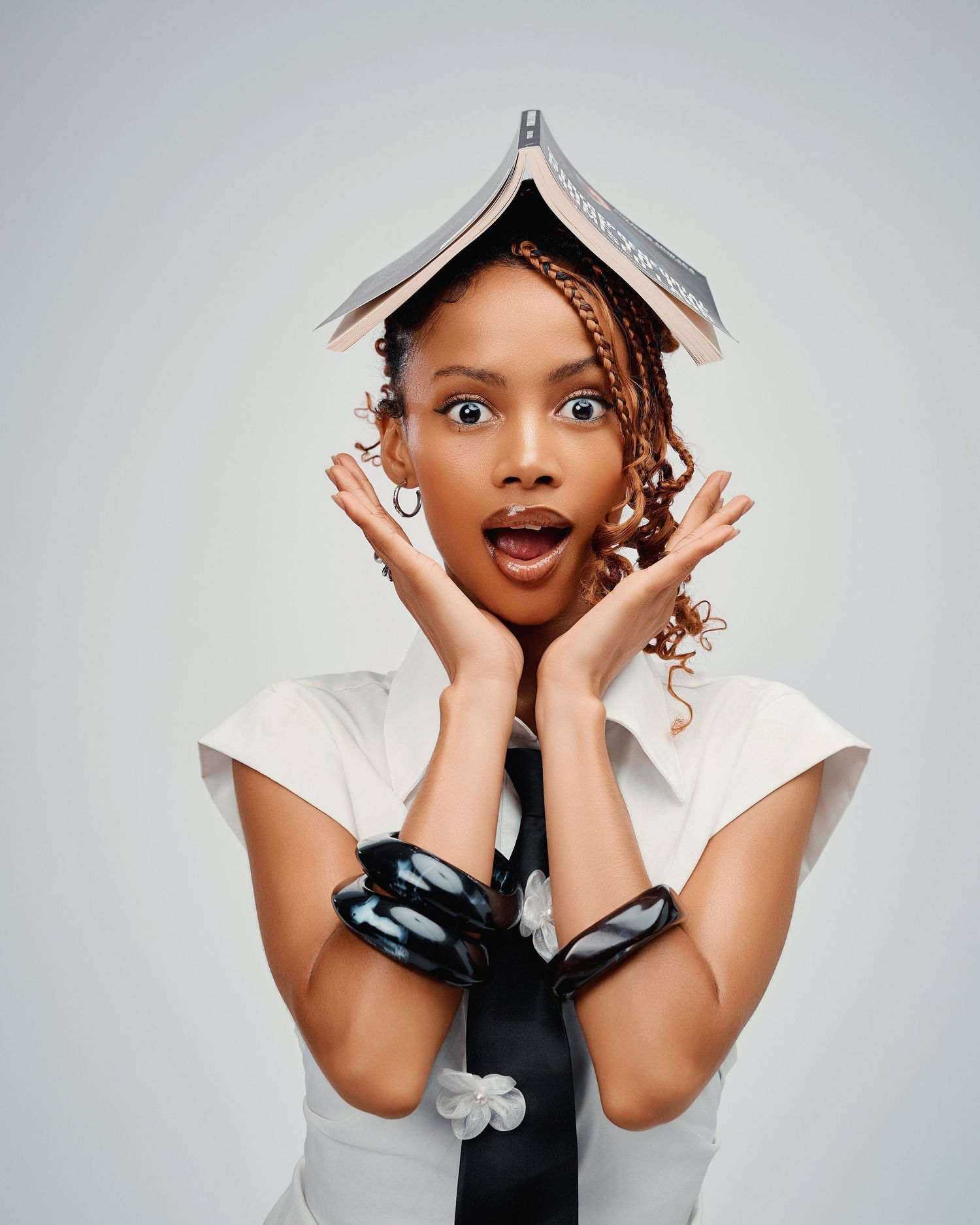 Surprised young woman with braids balancing an open book on her head, hands on cheeks, wearing white shirt and tie.