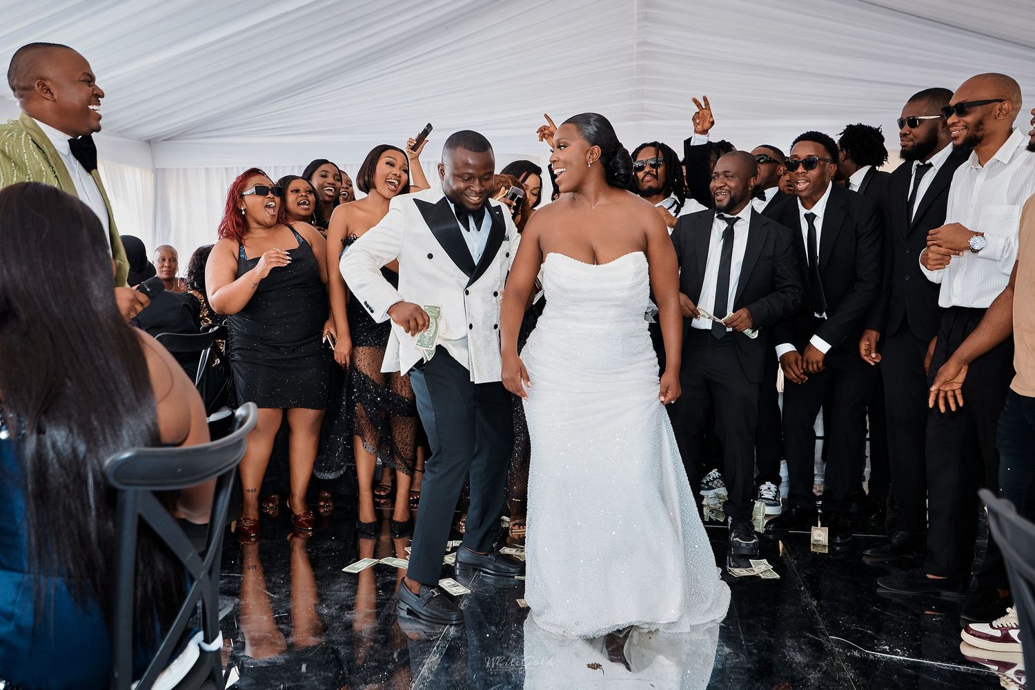 Bride in white strapless gown and groom in white tuxedo celebrating with guests at lively wedding reception.