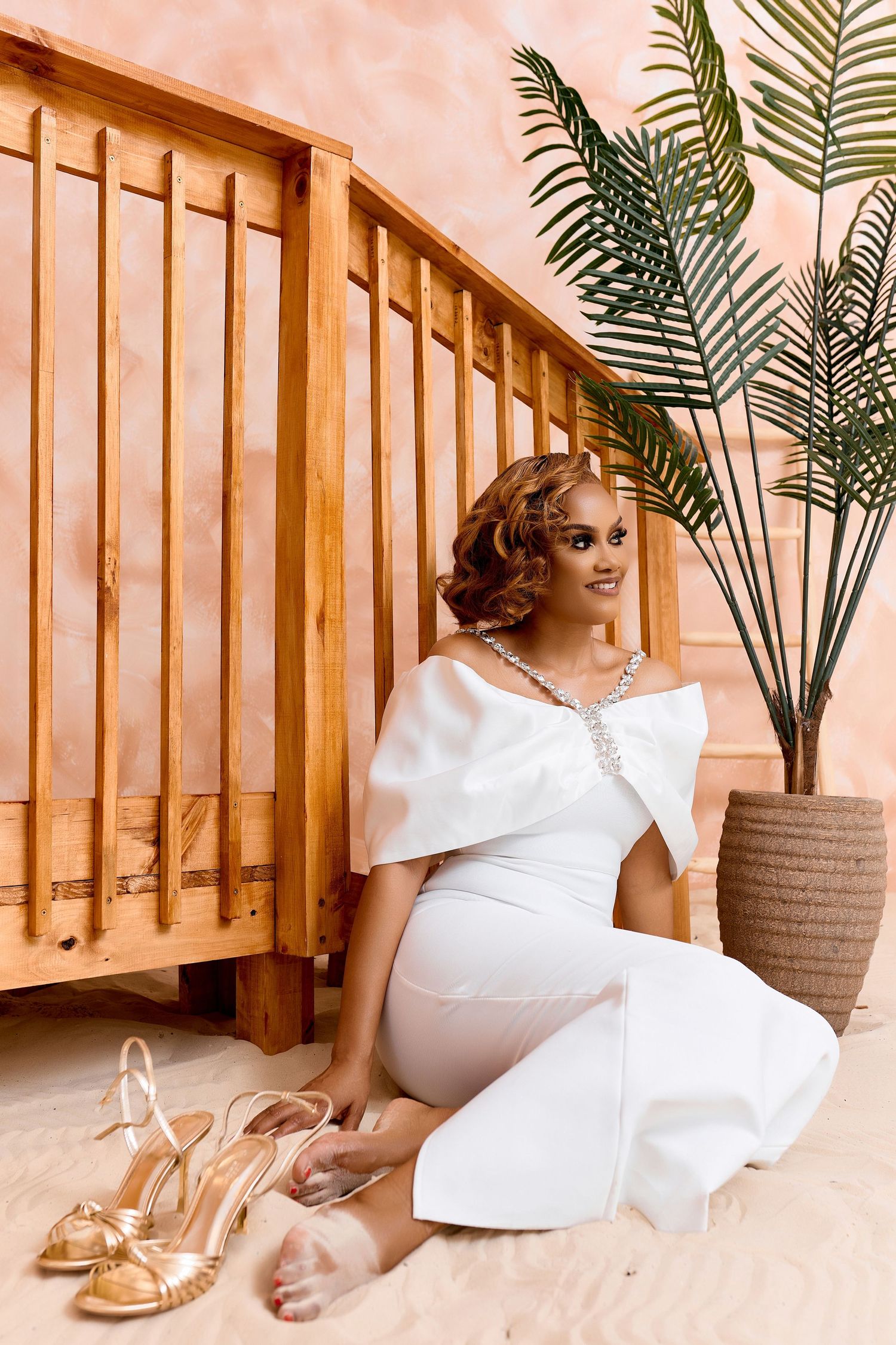 Stylish Black woman in white off-shoulder jumpsuit sits on sandy floor beside wooden staircase and palm plant.
