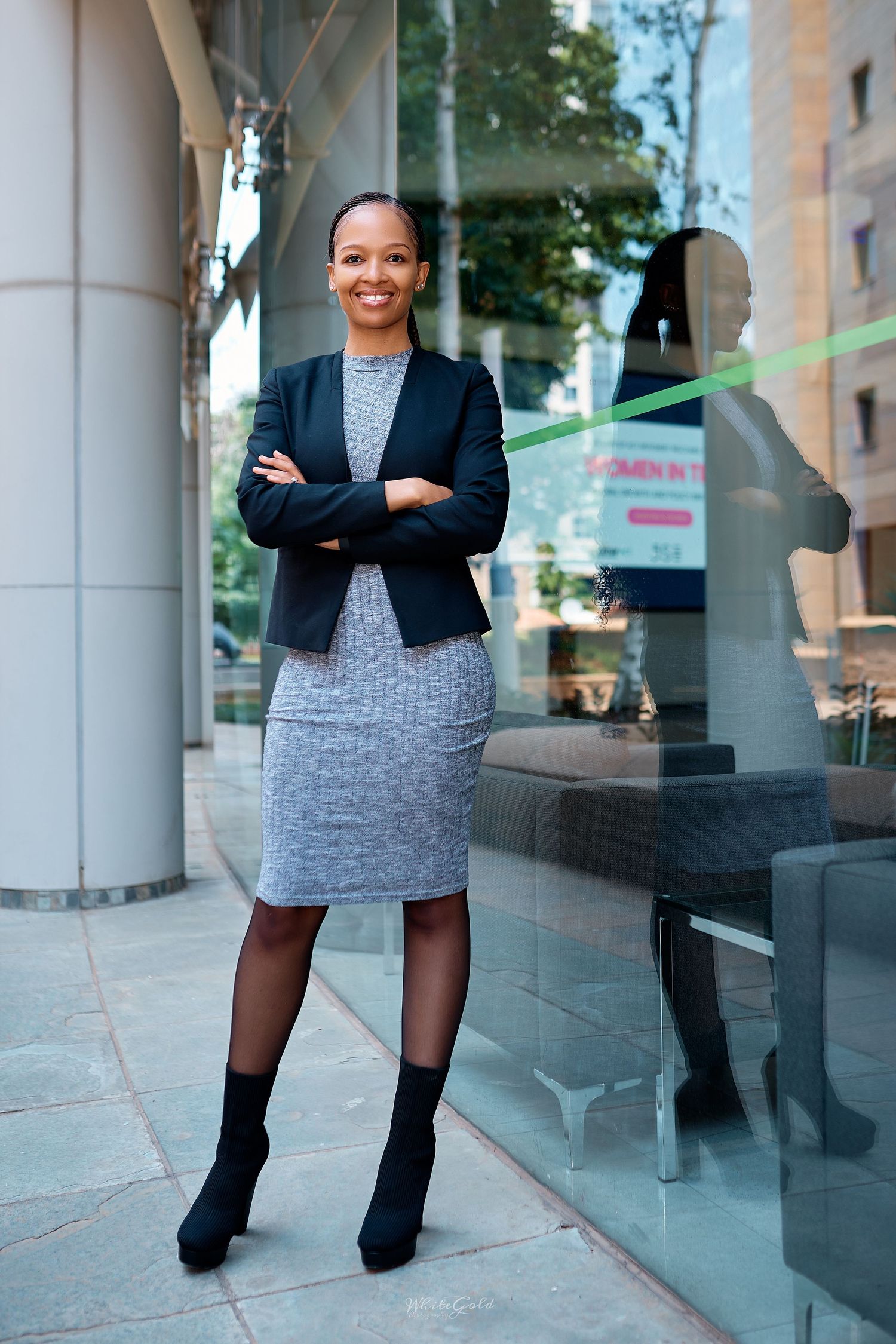 Smiling Black businesswoman in gray dress and blazer stands confidently outside a modern glass building.