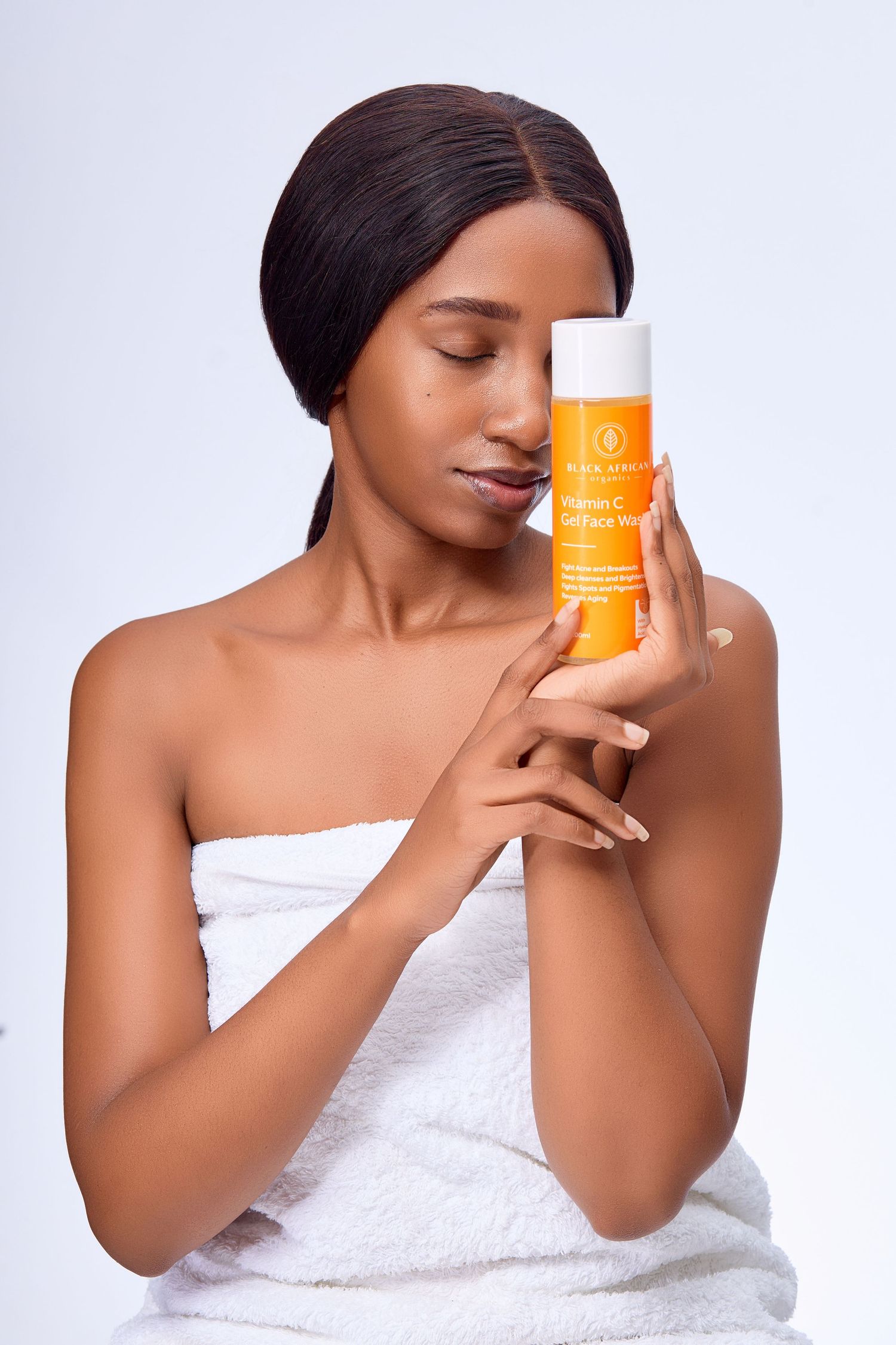 Woman in white towel holding orange Vitamin C gel face wash product against light background.