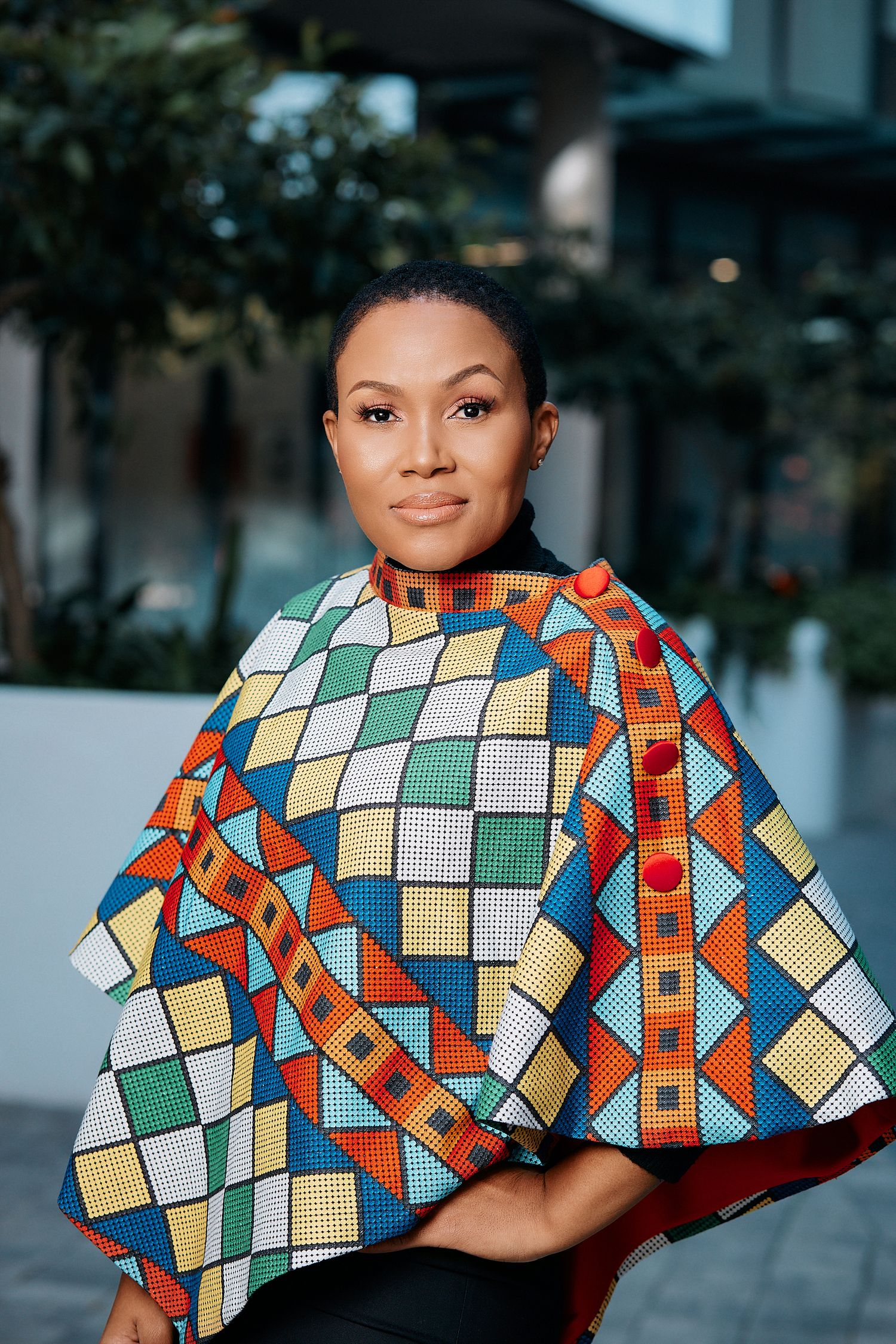 Woman wearing vibrant multicolor African geometric print poncho seated outdoors near building.