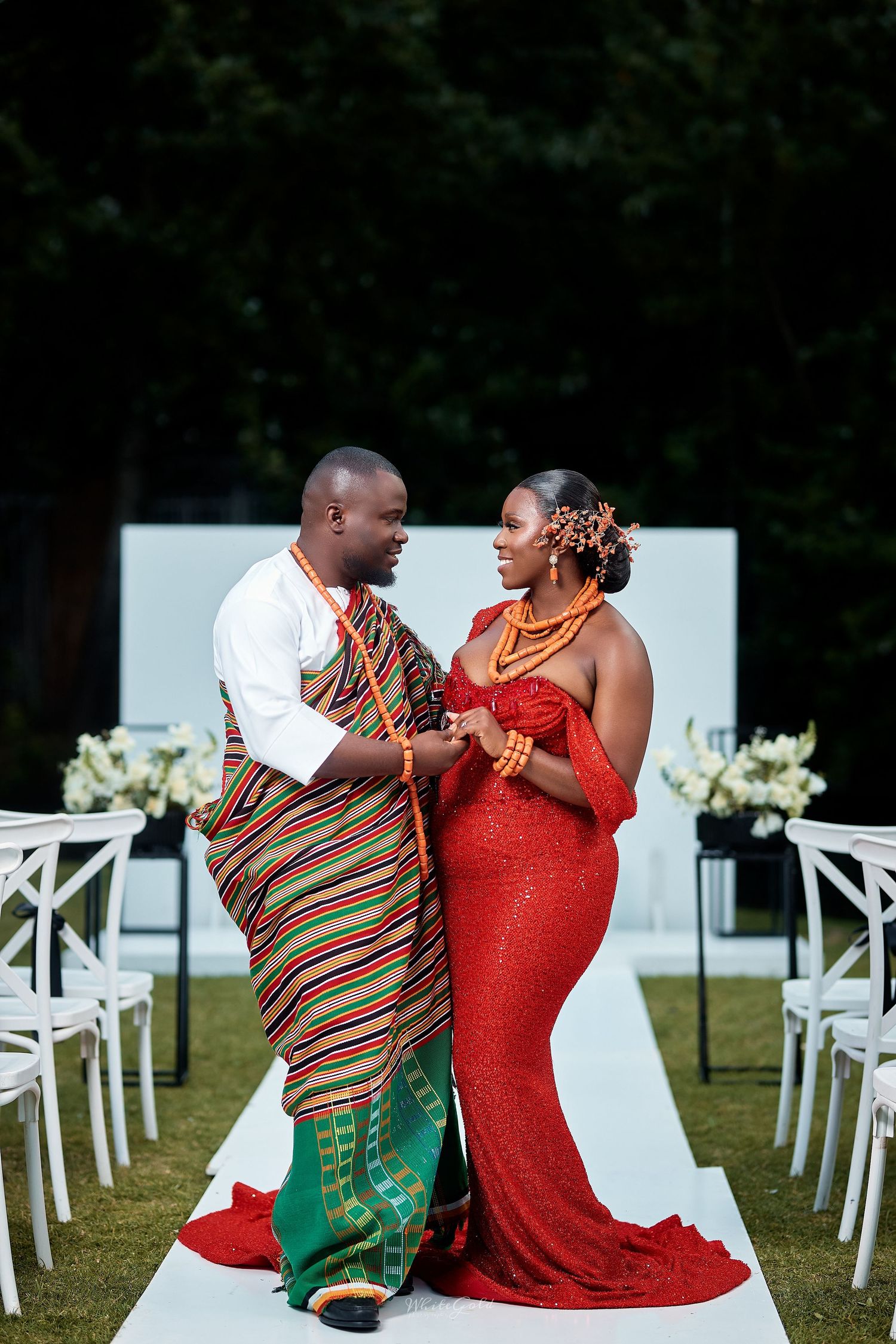 Couple in traditional Ghanaian kente and red attire standing at outdoor wedding aisle.