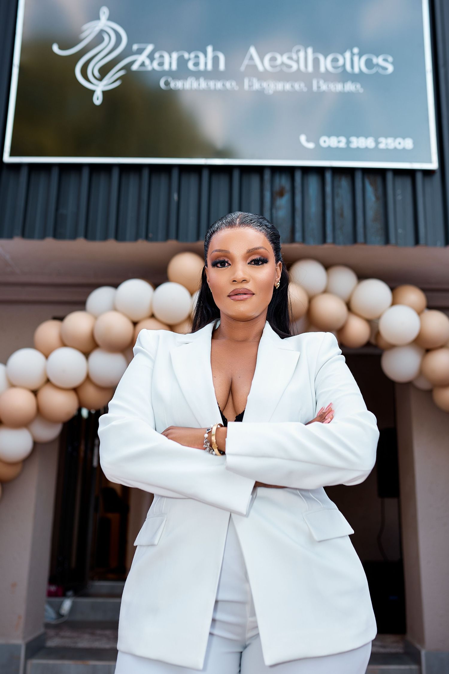 Confident woman in white blazer stands arms crossed before Zarah Aesthetics salon entrance with balloon decor.