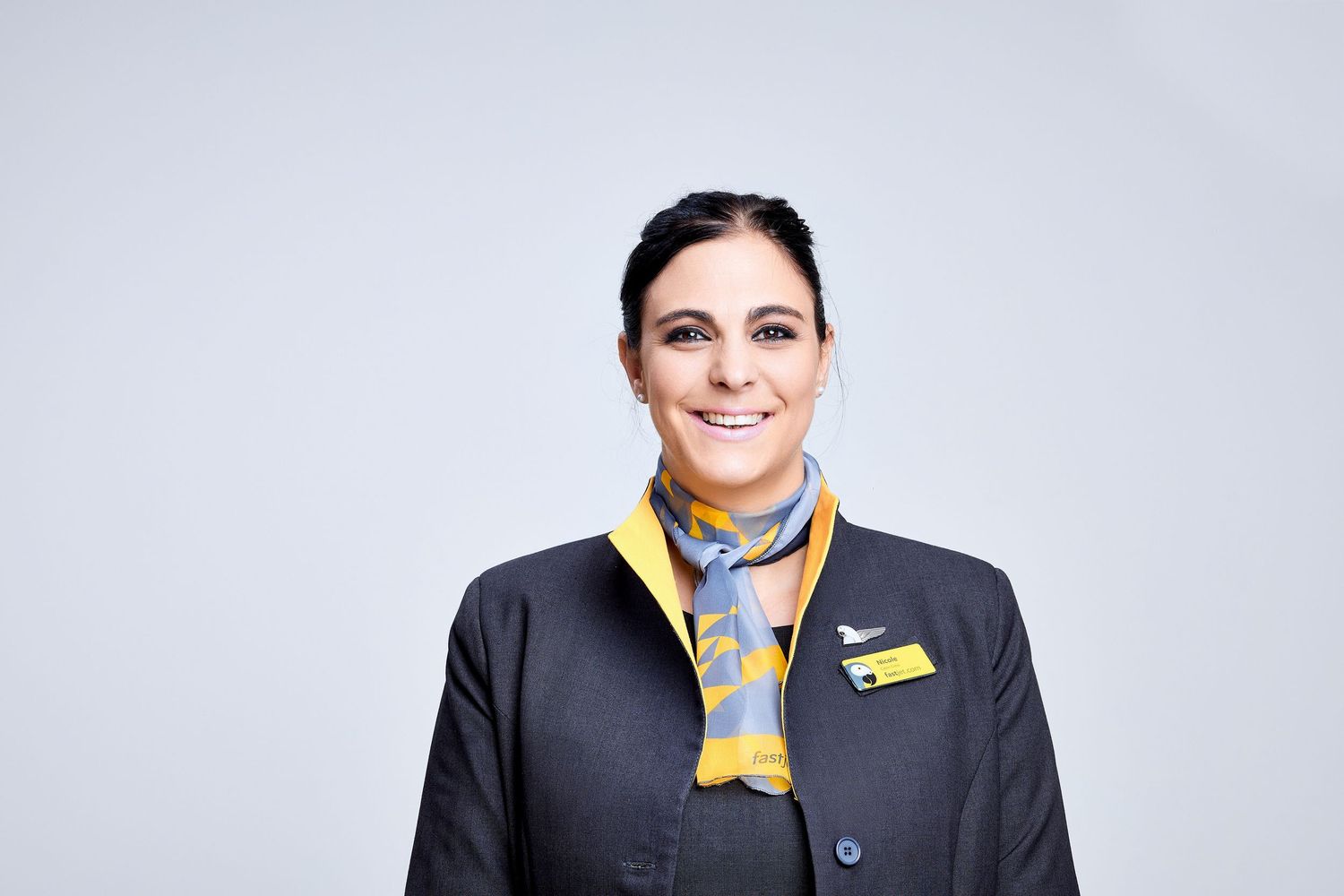 Smiling female flight attendant in dark uniform with yellow and blue scarf and airline badge on gray background.