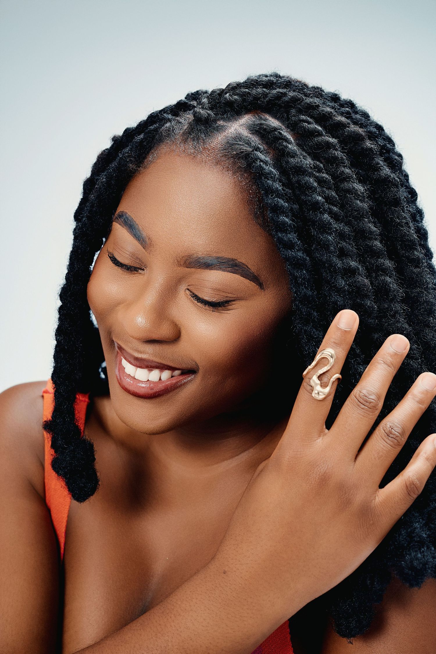 Smiling Black woman with twisted braids showcases gold statement ring against soft gray background.