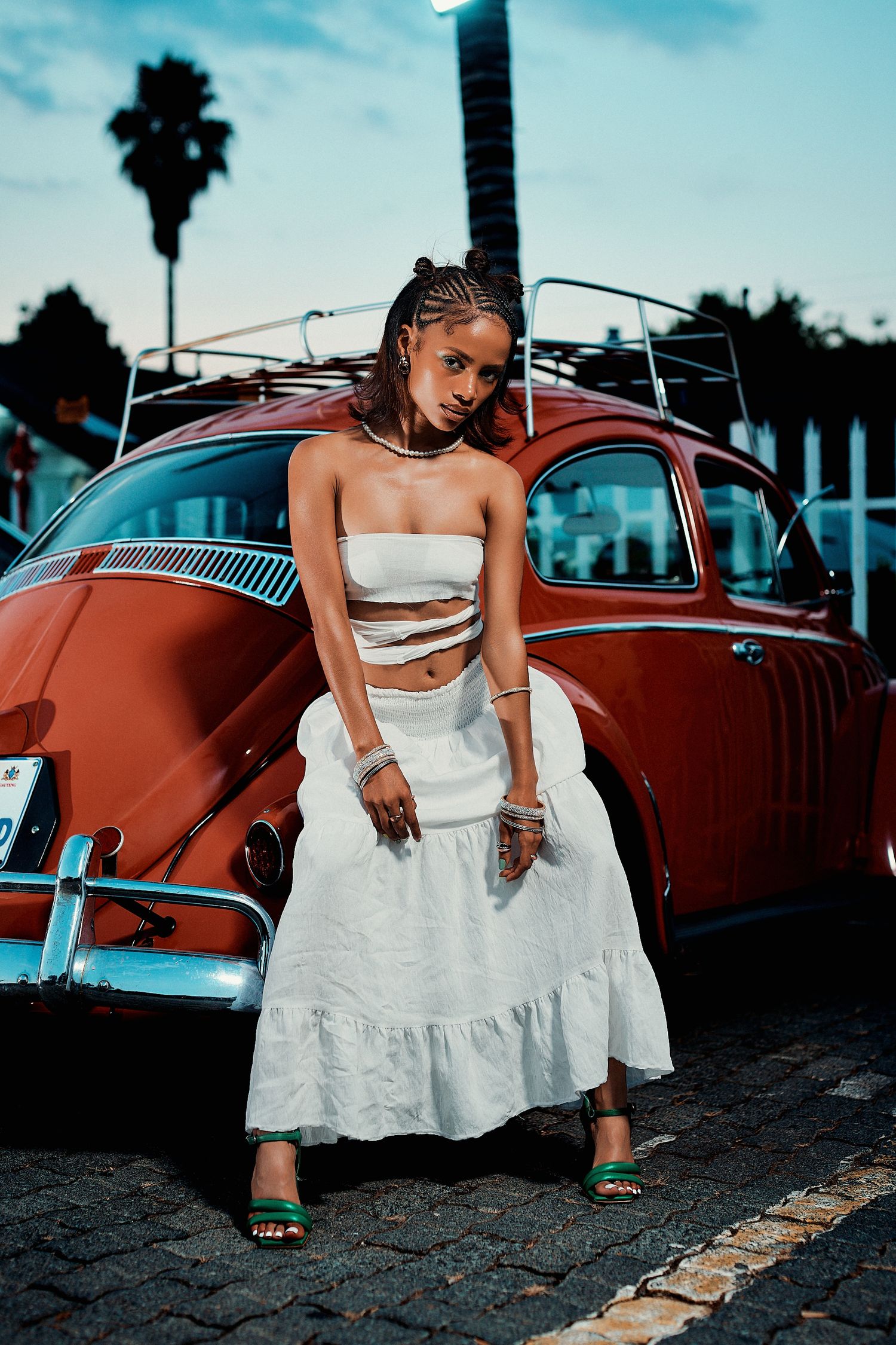 Young woman in white strapless crop top and maxi skirt stands beside a classic red Volkswagen Beetle at dusk.