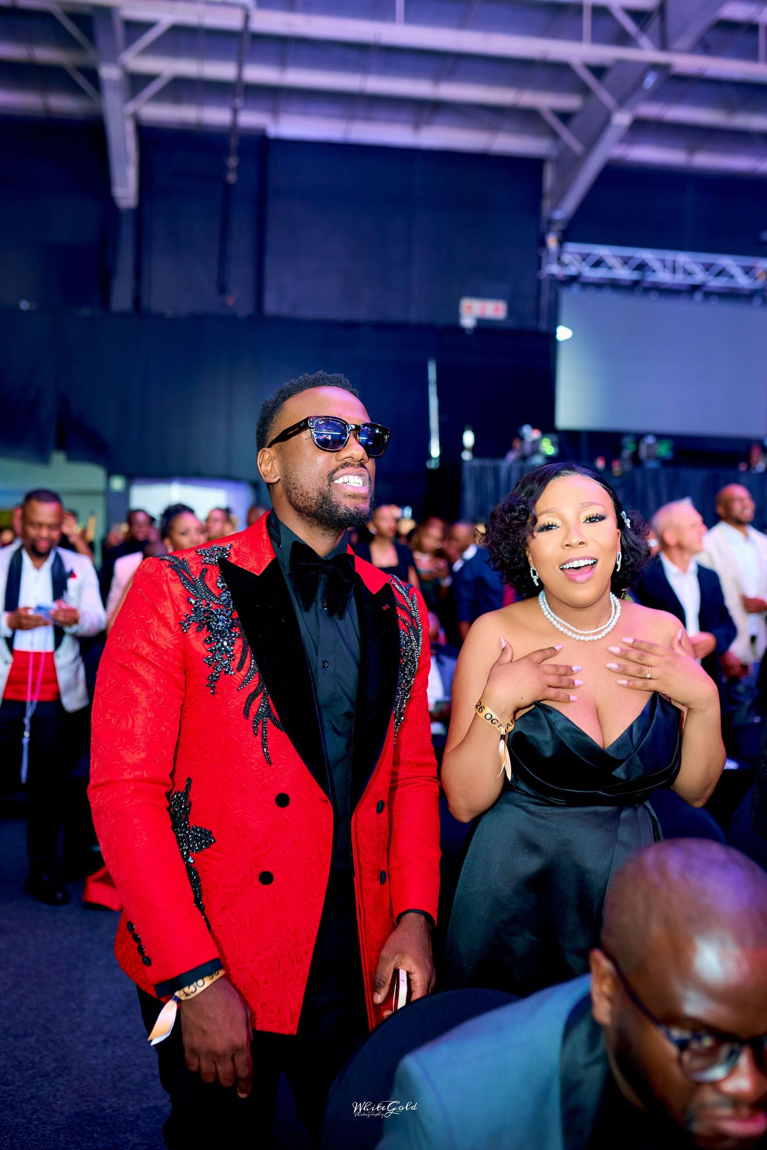 Stylish man in red embroidered blazer and sunglasses with woman in black dress at formal event.