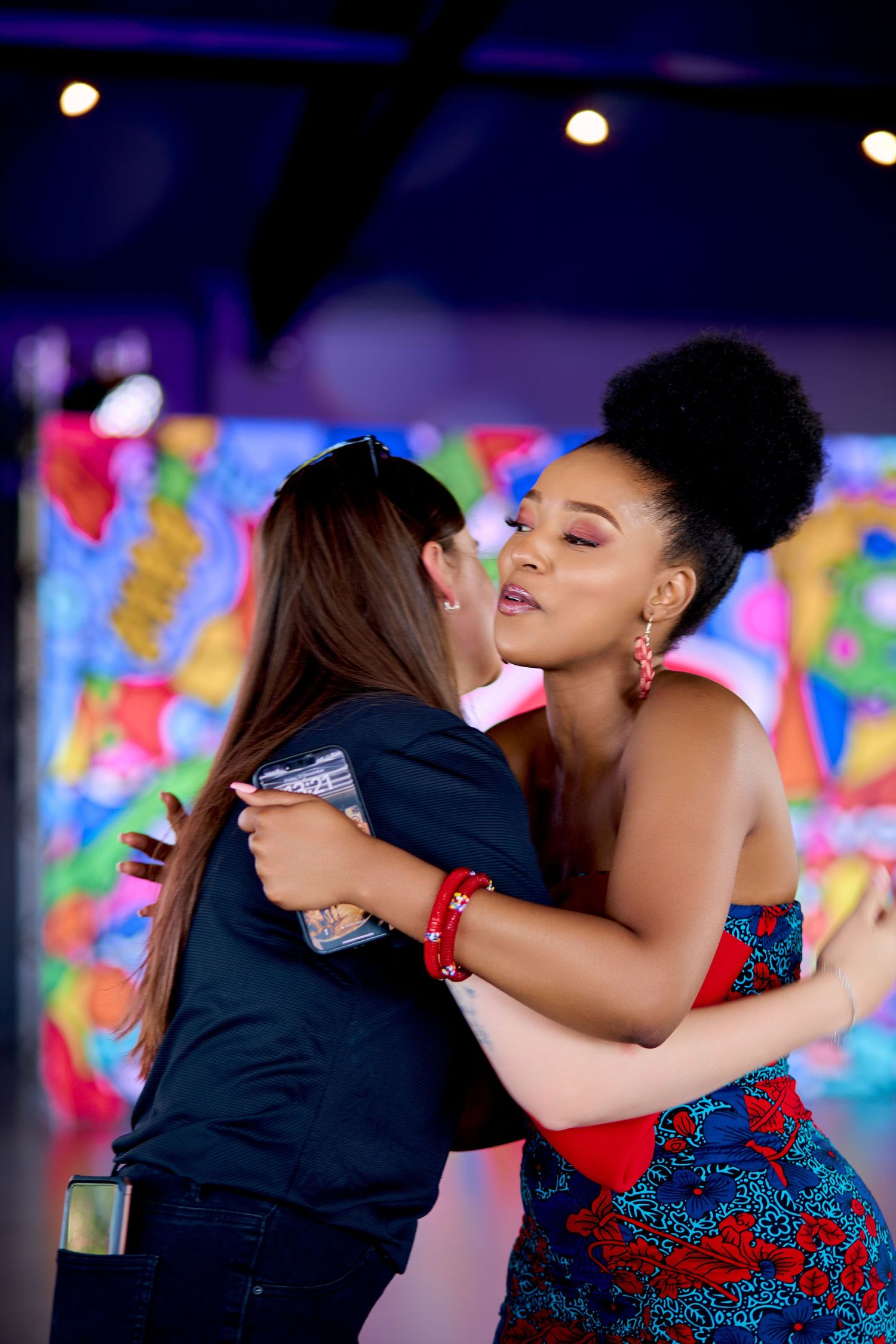Two women sharing a warm embrace at a colorful vibrant indoor event venue.
