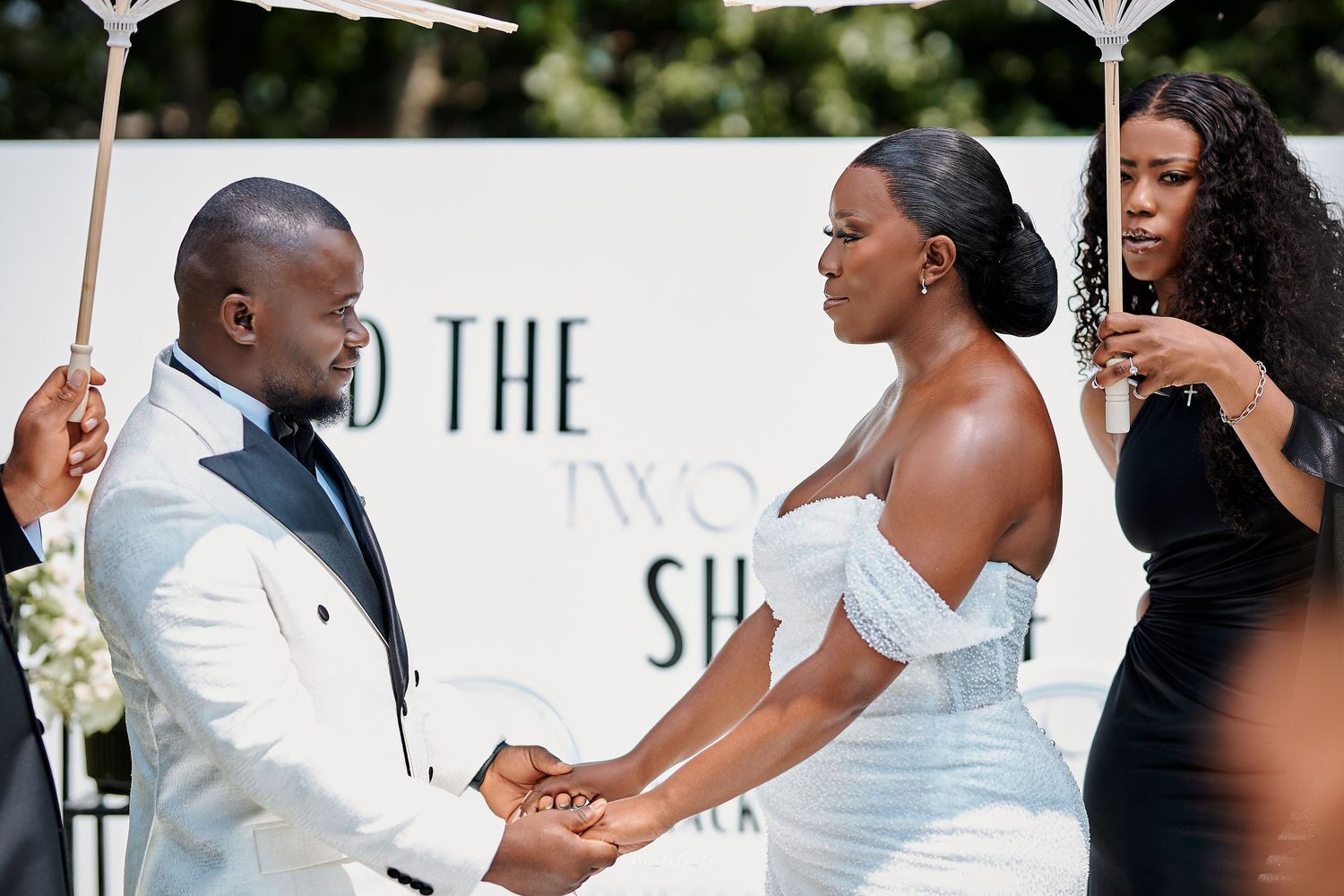 Groom in white tuxedo holding hands with bride in off-shoulder gown at outdoor wedding ceremony.
