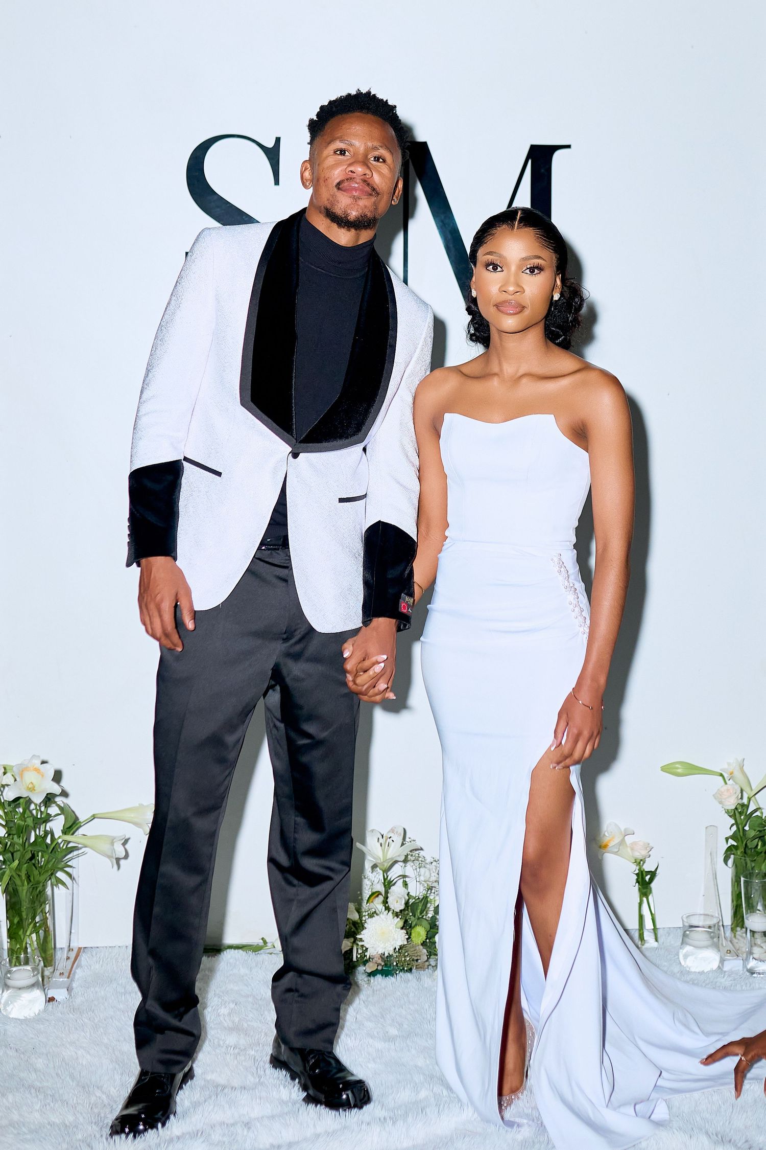 Elegant couple posing at formal event, man in white tuxedo, woman in white strapless slit gown.