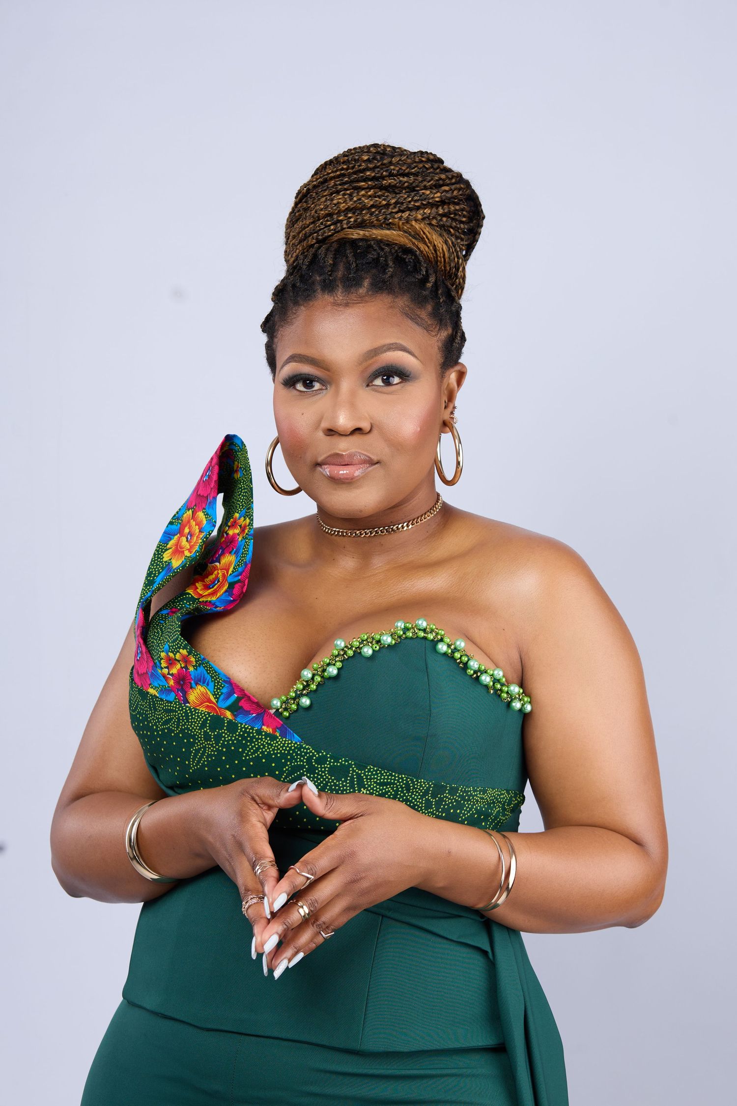 Woman in green strapless dress holding colorful heels, wearing gold jewelry and braided updo.
