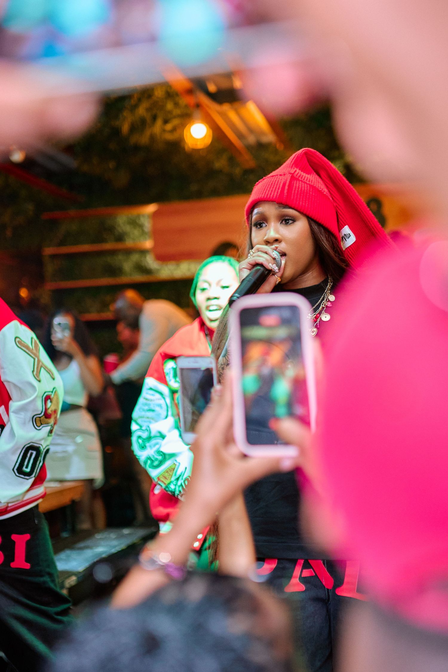 Female rapper in red beanie performs on stage with microphone as fans record with smartphones nearby.