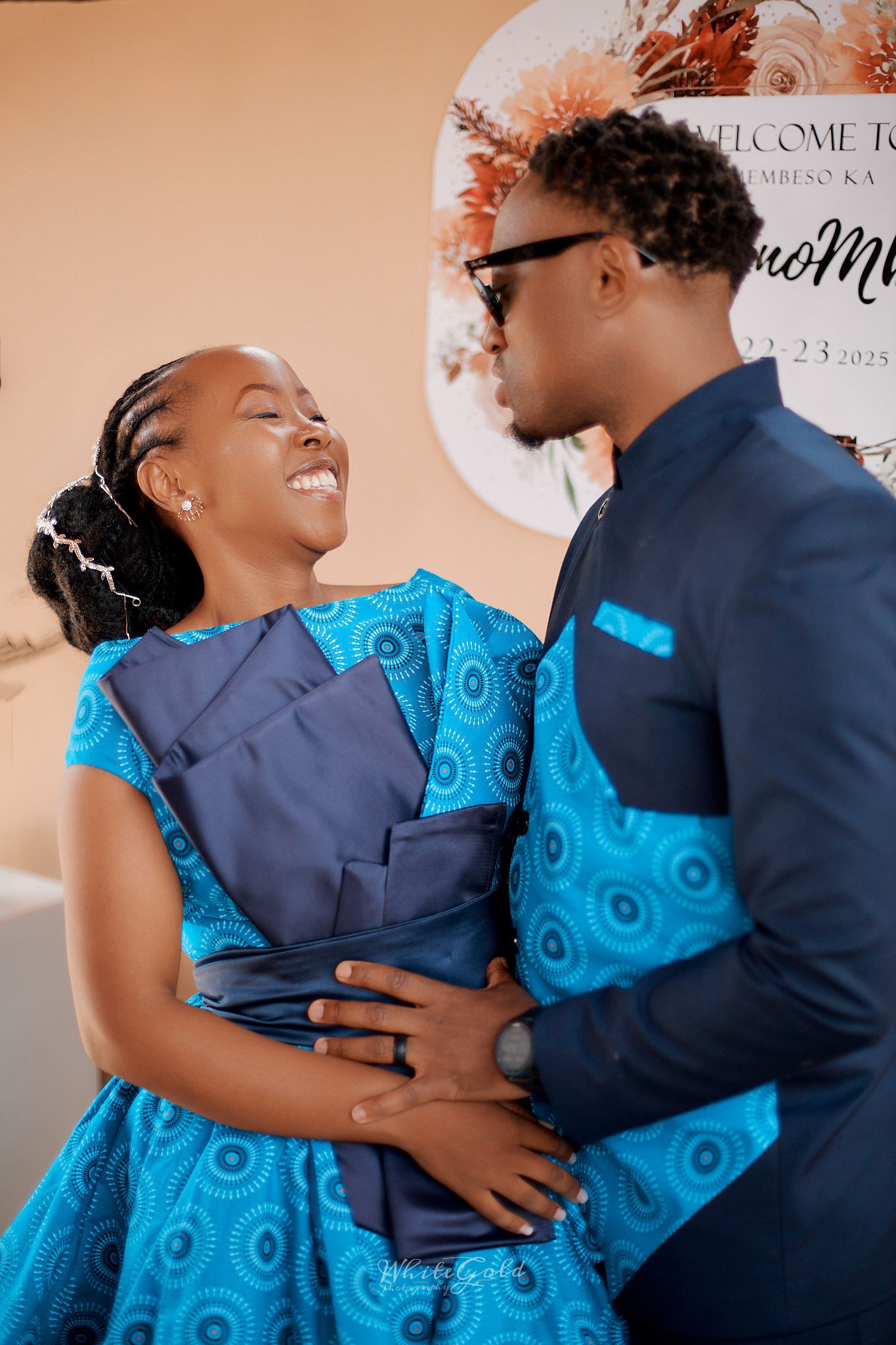 Couple in matching blue African print outfits smiling at each other at a wedding celebration event.