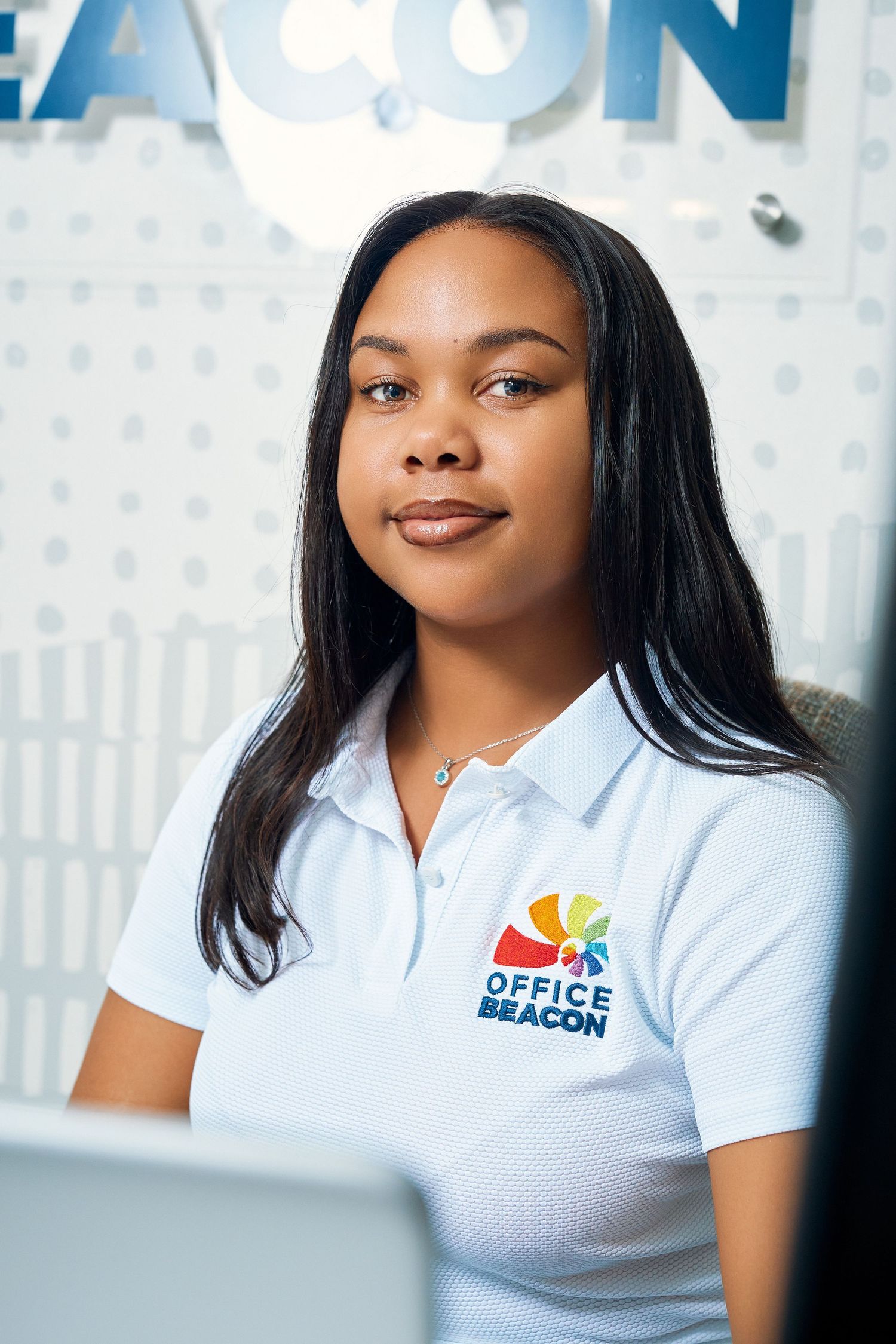Professional woman in Office Beacon white polo shirt smiling at workplace reception desk.