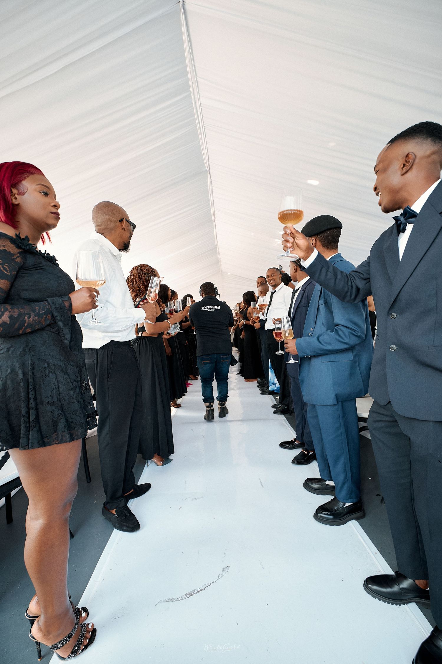 Elegantly dressed wedding guests in black attire raising champagne glasses in a toast inside a white tent.
