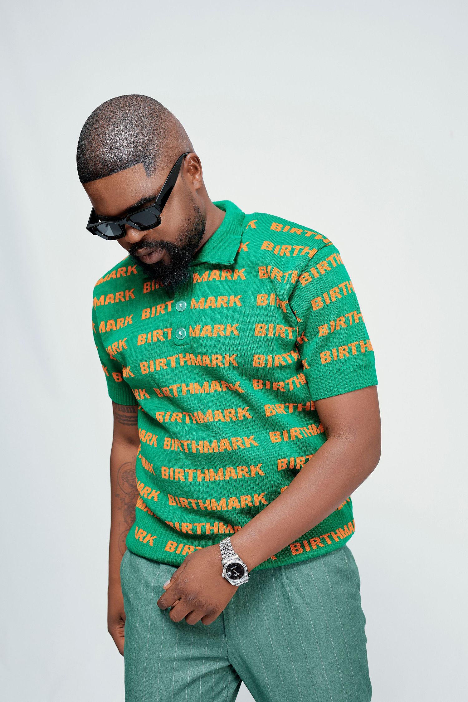 Man wearing green and orange Birthmark logo knit polo shirt with sunglasses looking down.