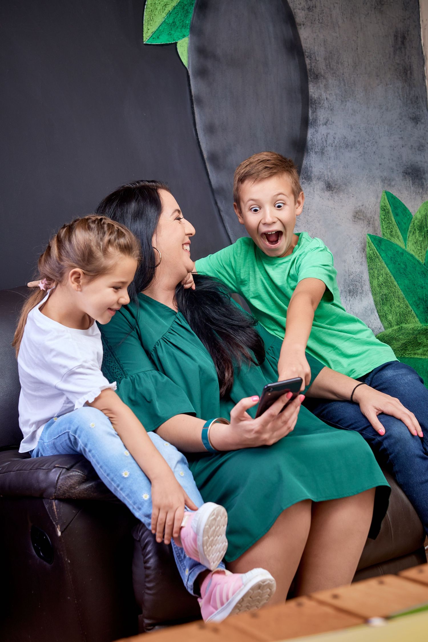 Happy mother in green dress sits on sofa with two excited children looking at a smartphone.