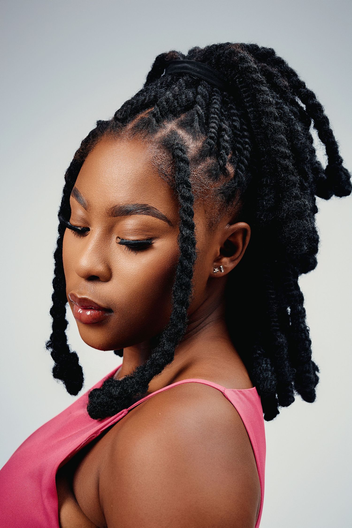 Profile view of a Black woman with elegant twisted locs updo hairstyle wearing a pink top on grey background.