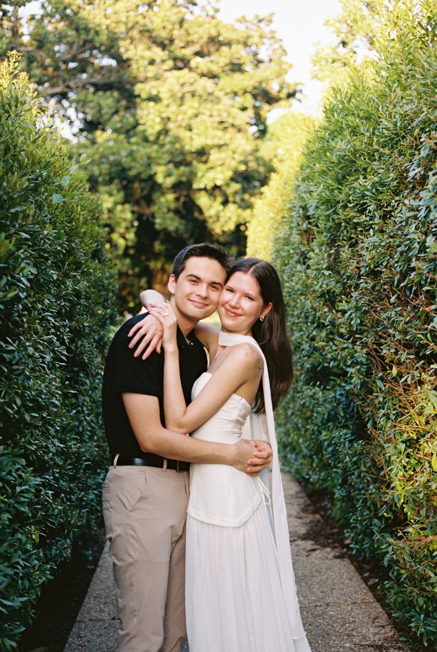 A smiling couple embraces in a lush garden pathway, woman in white dress and man in black shirt.