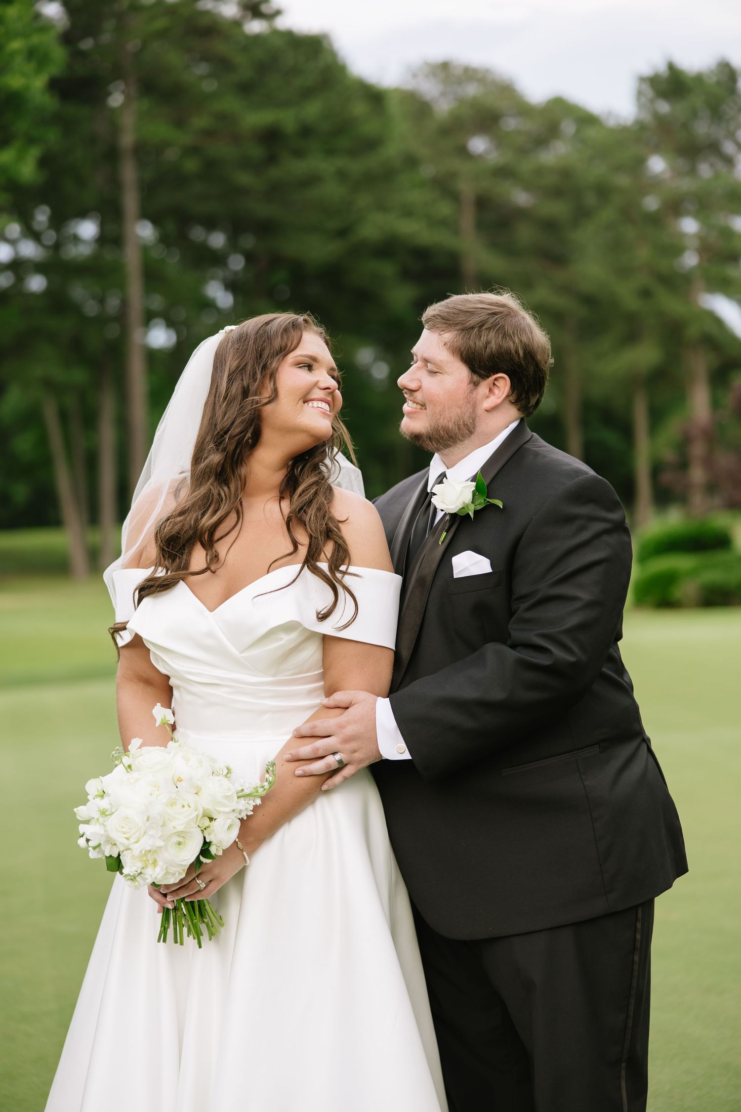 Bride in off-shoulder white gown holding white bouquet smiles at groom in black tuxedo outdoors on green lawn.